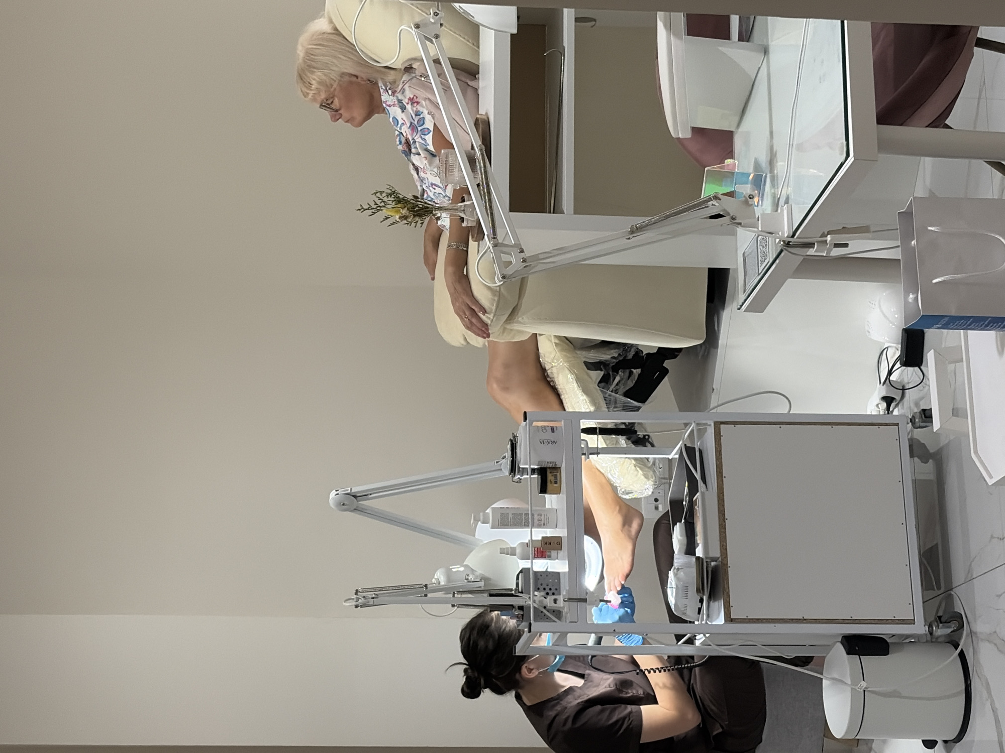 The image depicts a scene in a nail salon, likely located in Budva, Montenegro, during daylight hours. In the foreground, a technician, wearing a brown uniform and a light blue mask and gloves, is performing a pedicure on a client. The technician is seated and working on the client's feet with various tools. A white trolley cart holding a variety of nail care products, including bottles, jars, and a UV lamp, is positioned between the technician and the client.
The client, an elderly woman with blonde hair and glasses, is reclined in a large, comfortable-looking cream-colored chair. She is wearing a floral-patterned top. Beside her on a small wooden tray is a vase with a small arrangement of dried flowers and a glass of water.
In the background, a white wall forms the backdrop. To the right, another white table with a glass top is visible, holding a nail dryer and other items. A white shopping bag and a pink chair are also present in this area. The floor is tiled with a white and grey pattern. The overall environment suggests a clean and professional setting for nail services.