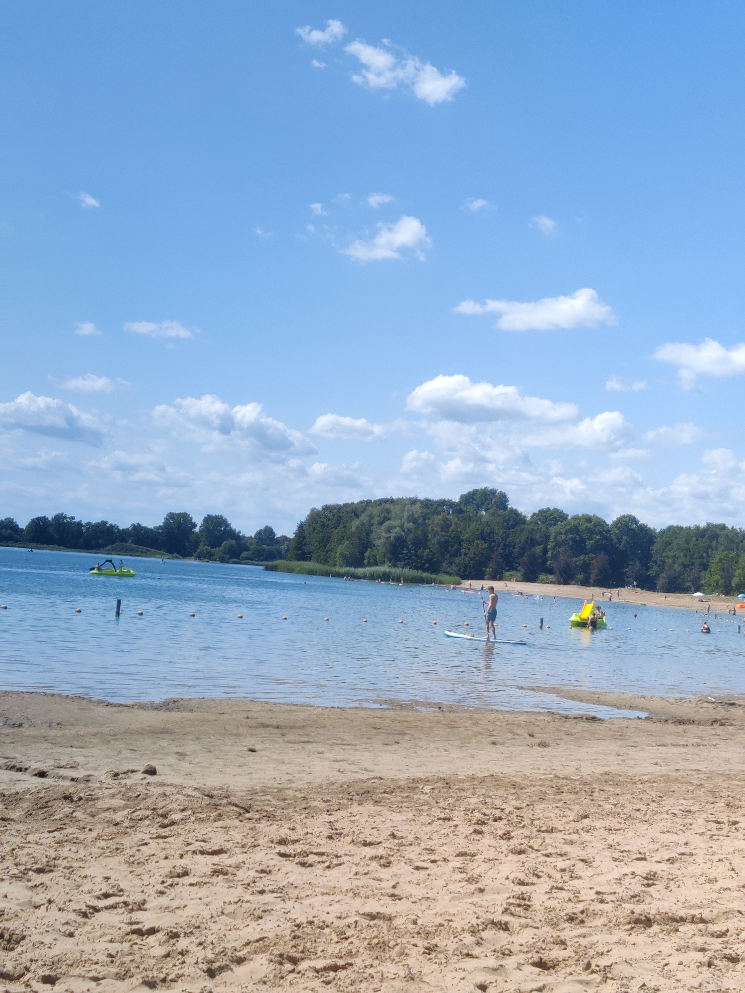 The image depicts a sunny day at a lake beach in Winterswijk, Netherlands. The foreground is a sandy beach with visible footprints and textured sand. The middle ground features a calm body of blue water with several people engaged in recreational activities. One person is standing on a paddleboard in the center of the frame. To the right, a yellow pedal boat with a slide is occupied by at least two individuals. Further in the distance, the water is dotted with buoys. The background consists of a lush, green tree line and a bright blue sky with scattered white clouds. The overall scene conveys a peaceful and summery atmosphere.