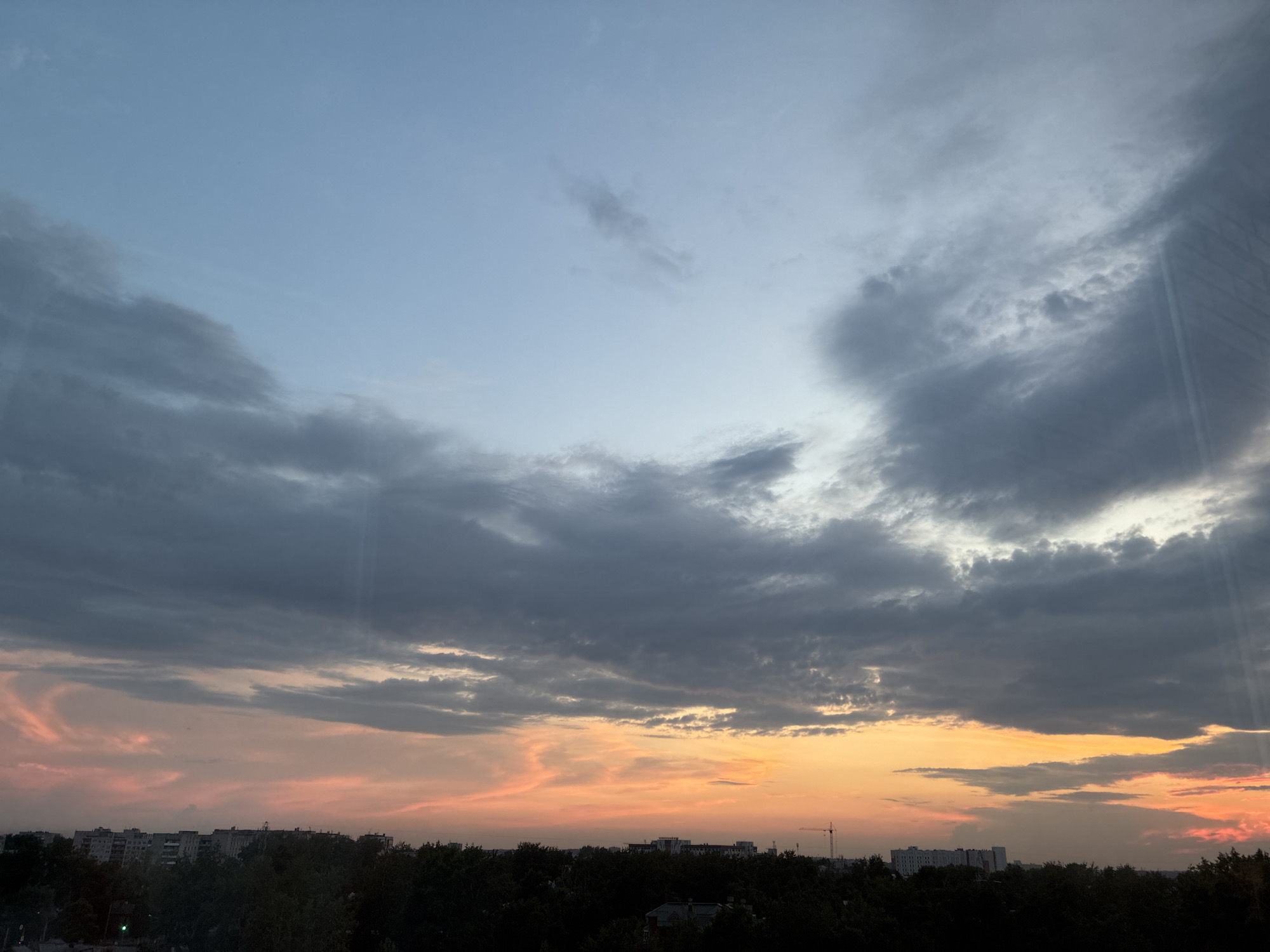 This image depicts a twilight sky over a cityscape. The sky transitions from a pale blue at the top to hues of orange and pink near the horizon, indicating sunset or sunrise. Dark, heavy clouds are present in the upper and middle portions of the sky, with some lighter, wispy clouds scattered throughout.
Below the sky, a dark silhouette of trees and buildings forms the lower part of the image. The buildings appear to be residential blocks, with some appearing taller than others. A construction crane is visible in the distance, to the right of the frame, against the colorful sky. The overall scene is one of a suburban or urban environment at dusk, with a dramatic sky as the focal point. There are no people or animals visible in the image. The presence of buildings and infrastructure suggests an urban setting, but the specific location is not identifiable from the image alone. The image is captured from an elevated viewpoint, possibly from a window or balcony.