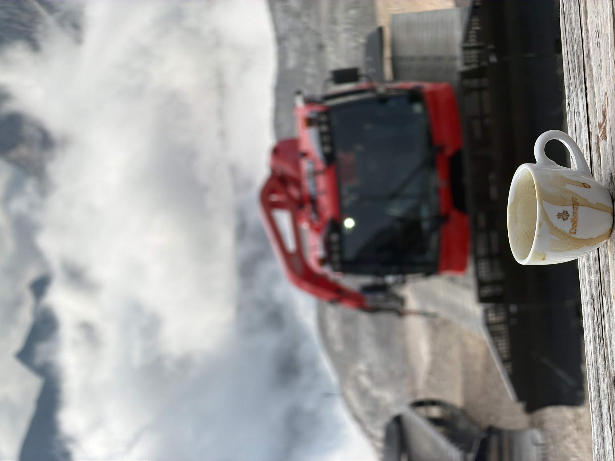 A white coffee cup with brown stains is in the foreground, placed on a weathered wooden surface. The cup has a small, gold logo with a crown. In the background, a large red vehicle, possibly a snow groomer or construction equipment, is visible. The vehicle has a black cabin with windows and red arm-like extensions. Beyond the vehicle, mountains are shrouded in clouds, creating a misty, atmospheric backdrop. The scene suggests a location in a mountainous region, likely Garmisch-Partenkirchen, Germany, given the presence of snow-related equipment and the mountainous terrain. There are no people visible in the image.