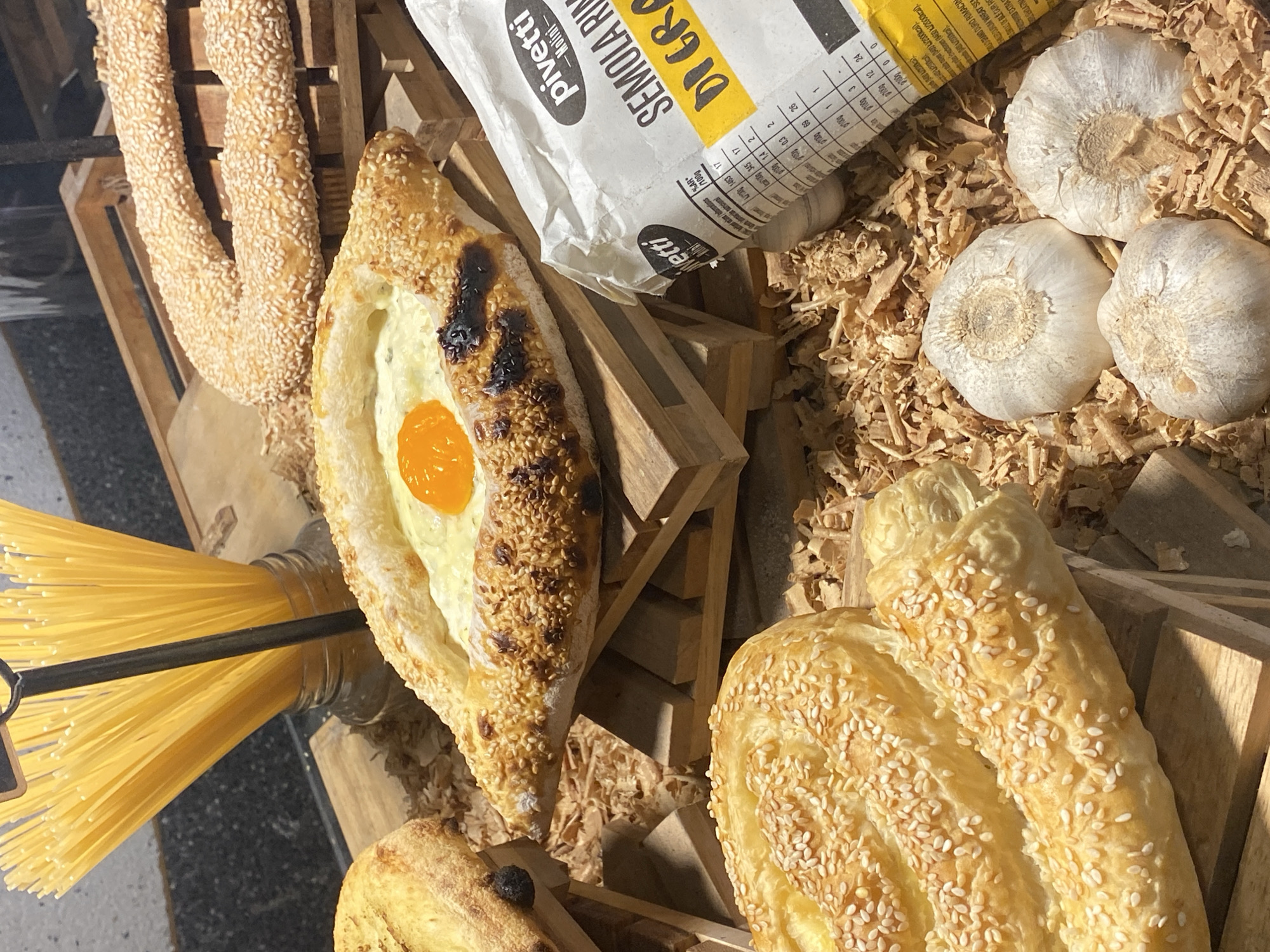 The image displays a rustic food presentation, likely a market stall or bakery display, located in Baan Chaweng, Thailand. Various baked goods and ingredients are arranged on wooden risers over a bed of light-brown wood shavings.
In the central foreground, a boat-shaped pastry, consistent with Khachapuri Adjaruli, is prominent. It features a golden-brown crust, generously sprinkled with sesame seeds, and exhibits some darker, charred areas. The central indentation holds a creamy white filling, likely cheese, topped with a single, vivid orange egg yolk.
To the upper right of this pastry, a large, elongated, and twisted bread item, heavily coated in sesame seeds, is visible. This item rests on a dark wooden crate. A similar, coiled or folded pastry, also covered in sesame seeds, occupies the bottom left portion of the frame. Another smaller, light-colored bread item is partially visible on the far left.
Behind the boat-shaped pastry, a clear glass jar is filled with numerous strands of uncooked yellow spaghetti, standing vertically. A slender black metal rod, possibly part of a display stand, is positioned behind the spaghetti jar.
In the lower right section of the image, a white paper bag, angled towards the viewer, displays partial text including "pivetti," "Molini," "SEMOLA RIN," and "DI GRAS," indicating it is a semolina flour product. Adjacent to this bag, nestled within the wood shavings, are three distinct, whole garlic bulbs.
The food items are presented on dark wooden crates and slats, creating elevated platforms. The base of the display is covered with a layer of fine, light-brown wood shavings or shredded wood. The background is dark and out of focus, suggesting an indoor setting.