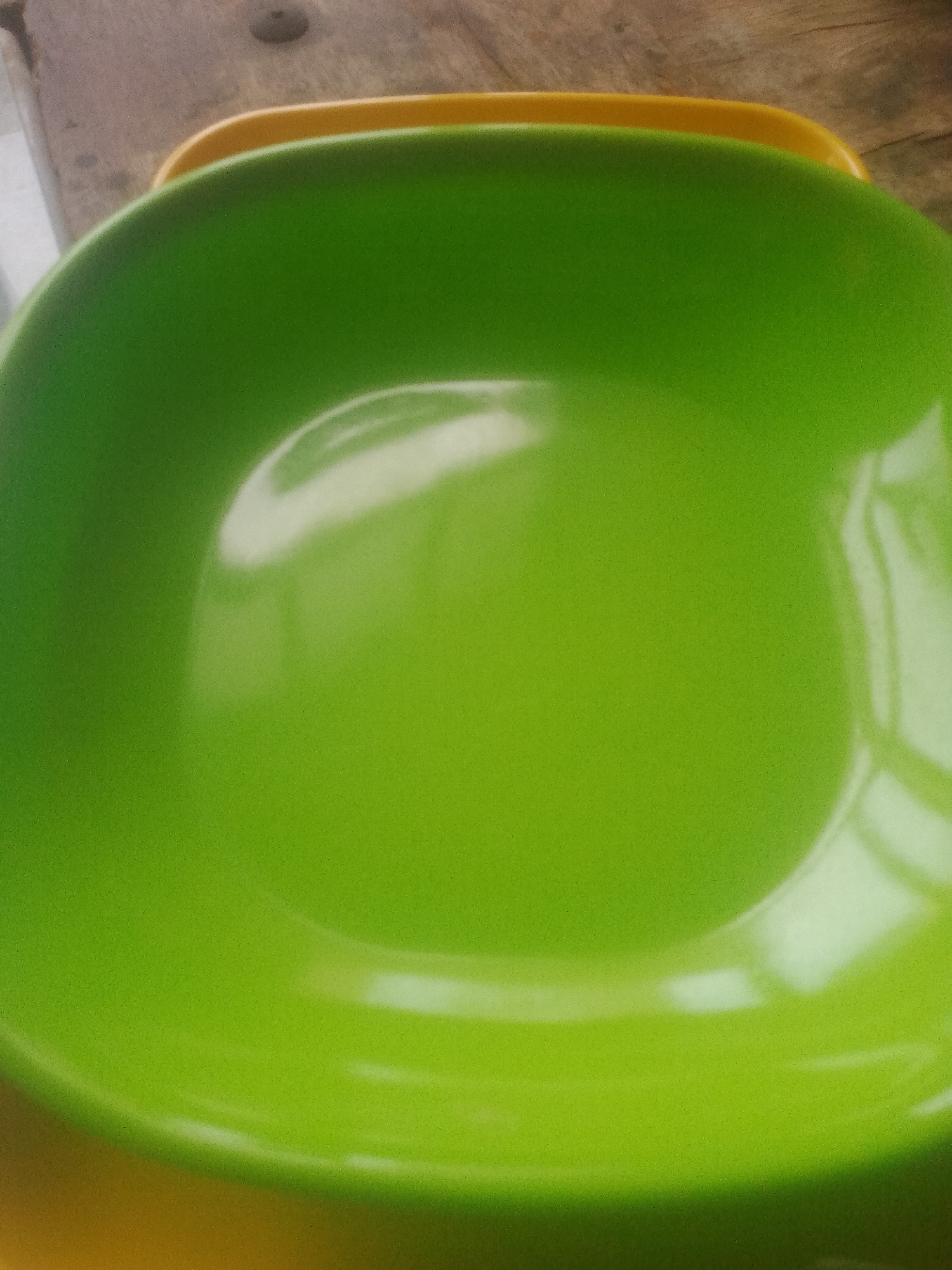 The image shows a close-up of two nesting bowls. The larger bowl is lime green and the smaller bowl beneath it appears to be yellow. The bowls are resting on a wooden surface, possibly a table. The lighting suggests it is daytime and the reflections on the green bowl indicate it is smooth and possibly made of plastic or ceramic. There are no people or any other objects visible in the frame. The image does not provide any information to associate it with Jauro Yinu, Nigeria or any other specific location.
