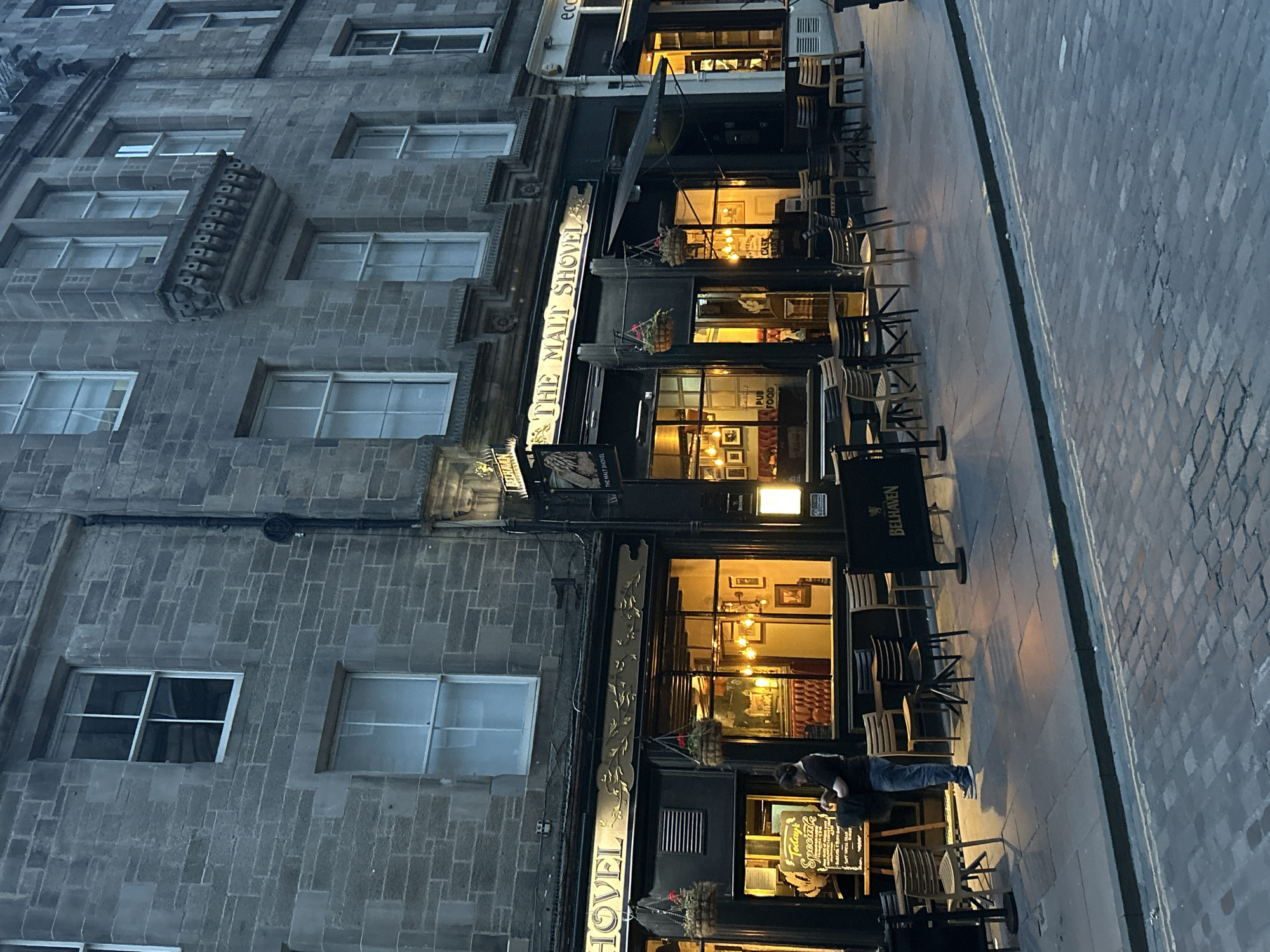 The image shows the exterior of "The Malt Shovel" pub, located on a cobblestone street in the City of Edinburgh, United Kingdom. The pub has a traditional stone facade and large windows that reveal a warmly lit interior. Outside the pub, several tables and chairs are set up on the sidewalk, some of which are under a black awning. A person in jeans and a dark t-shirt is standing by a "Today's Specials" blackboard outside the pub, looking down at a bag they are holding. The street in front of the pub is paved with cobblestones, and there are double yellow lines painted on the road. The building appears to be made of grey stone blocks. The sky is overcast, suggesting it is either early morning or late evening.