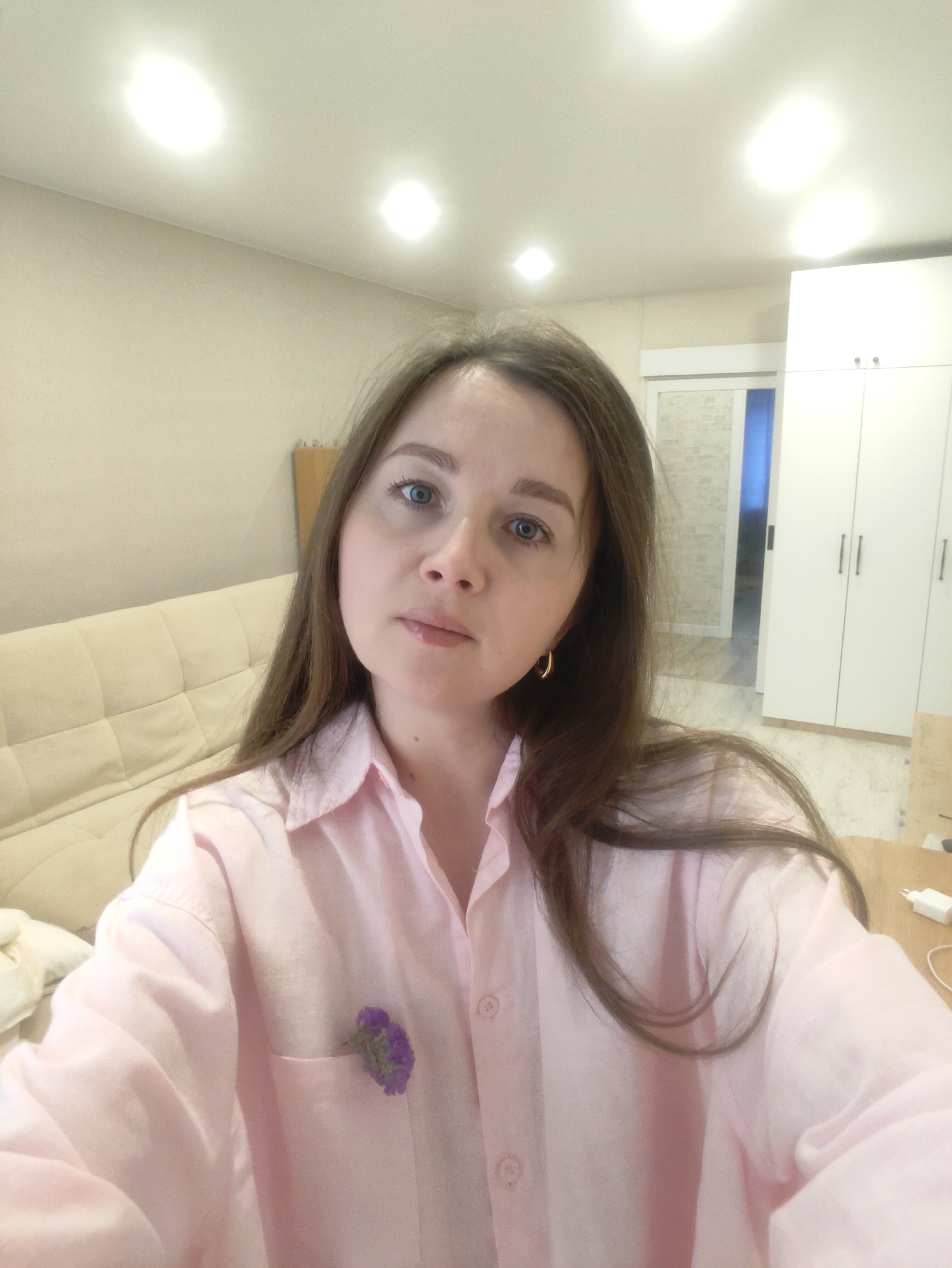 A woman is indoors, likely in an apartment in an unknown city. She has light skin, blue eyes, and brown hair. She wears a light pink button-down shirt with a small purple flower in the pocket. Behind her is a light-colored couch, white cabinets, and a doorway leading to another room. The ceiling has several recessed lights.