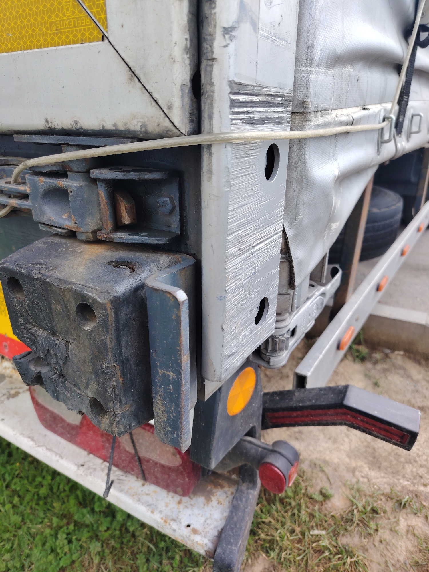 A close-up, eye-level, outdoor shot shows the rear corner of a large, weathered white trailer with a grey tarpaulin cover. The trailer has a heavy-duty, dark metal mechanism attached to its corner, featuring multiple bolts and interlocking parts. A thin, pale yellow rope is threaded through a hole in this mechanism and extends towards the right. To the right of the main mechanism, a black plastic component with red reflectors is mounted, along with a small red circular light. Part of a tire and a metal rail with orange reflectors are visible further back and to the right, suggesting the trailer is parked on a gravel or dirt surface with some dry grass. The overall impression is one of a functional, industrial vehicle in a practical, possibly rural or industrial, setting. The image was taken in Khorugvino, Russia.