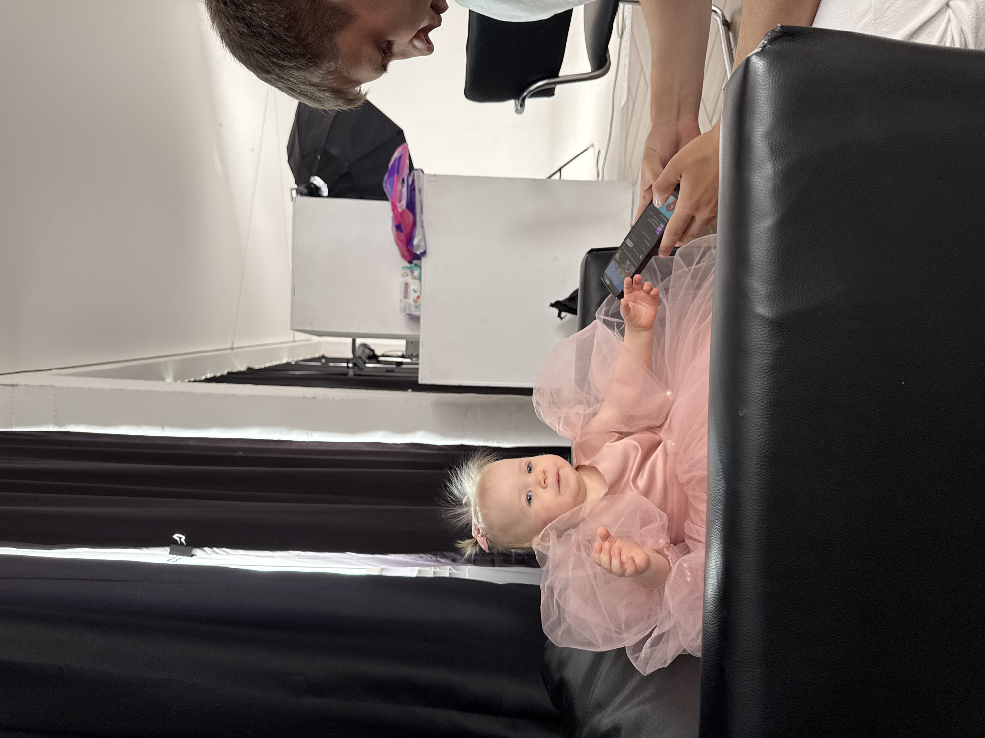 A young child wearing a pink dress is lying on a black surface. A person stands above the child, holding a smartphone. The room also contains a white table, an umbrella, and a chair. The location is Chaikovsky, Russia.