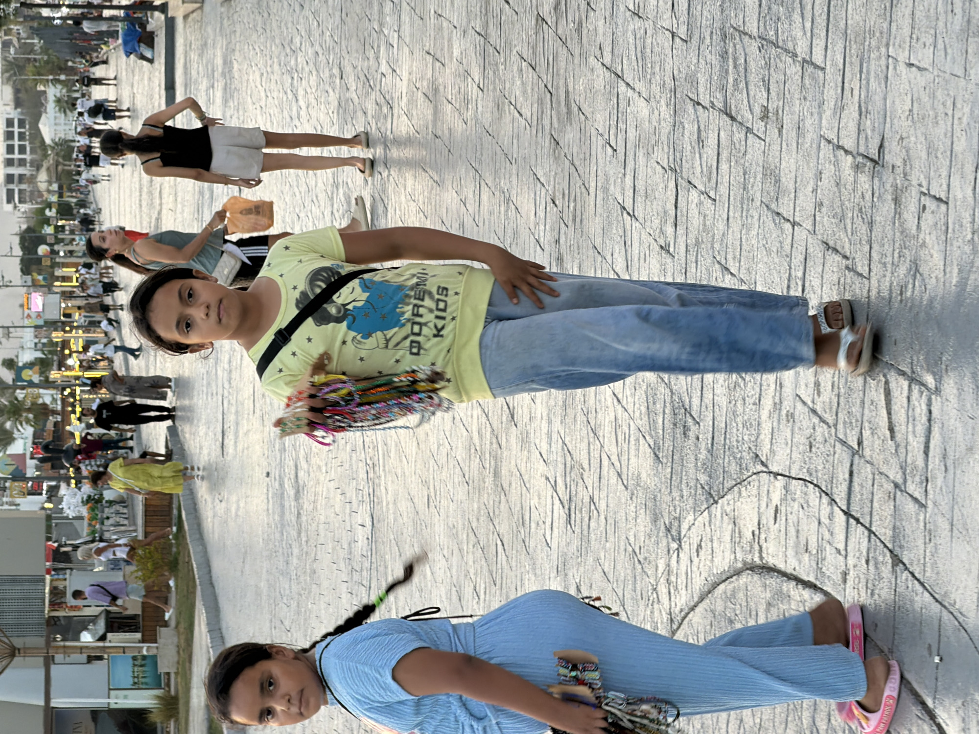 The image depicts a pedestrian walkway in Sharm El Sheikh, Egypt. In the foreground, two young girls are walking. The girl on the left is wearing a light blue textured tracksuit and pink flip-flops. She has a dark braid that extends to her left. She is carrying a small purse with colorful beaded items hanging from it. The girl on the right is wearing a yellow t-shirt with a graphic print and light blue jeans, and silver sandals. She is carrying a bundle of colorful beaded bracelets in her right hand. In the mid-ground, several other pedestrians are visible, including women in dresses and men in casual clothing. The walkway is paved with textured gray stones. In the background, buildings and palm trees are visible, suggesting an urban environment. The lighting suggests it is late afternoon or early evening.