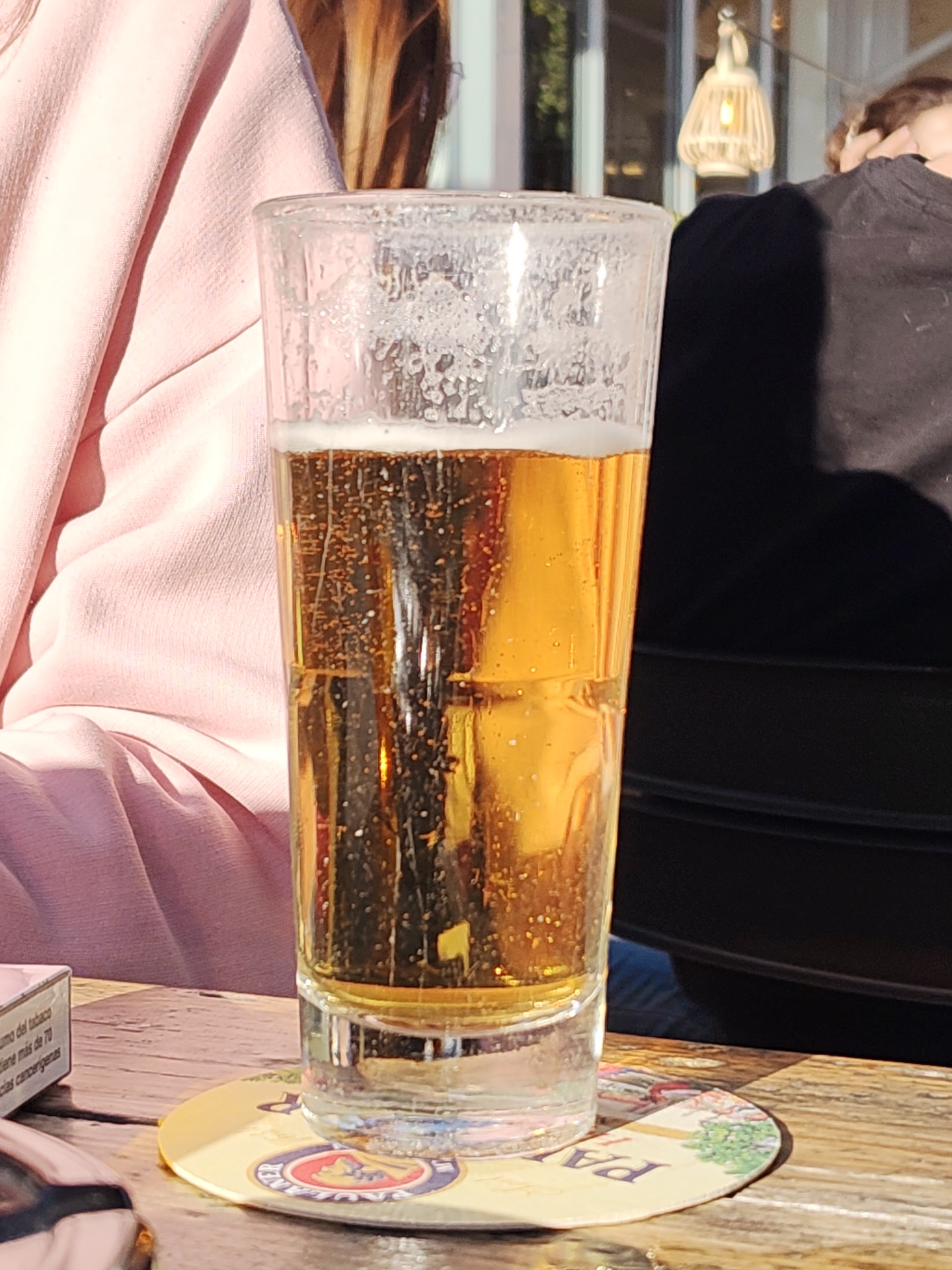 The image shows a glass of beer on a coaster that sits on a wooden table. The glass is almost full, with a frothy head. A person wearing a pink sweater is visible to the left of the glass, while another person in black clothing is partially visible to the right. A pack of cigarettes is present on the table to the left of the glass. The setting appears to be outdoors, possibly a restaurant or cafe. The coaster features the brand name "Paulaner," indicating the type of beer.
Given the prevalence of the Paulaner beer brand and the general style of outdoor dining common in tourist areas, the location is likely in Dénia, Spain, a coastal city with a significant German expatriate population. This suggests the presence of establishments serving popular German beers like Paulaner.