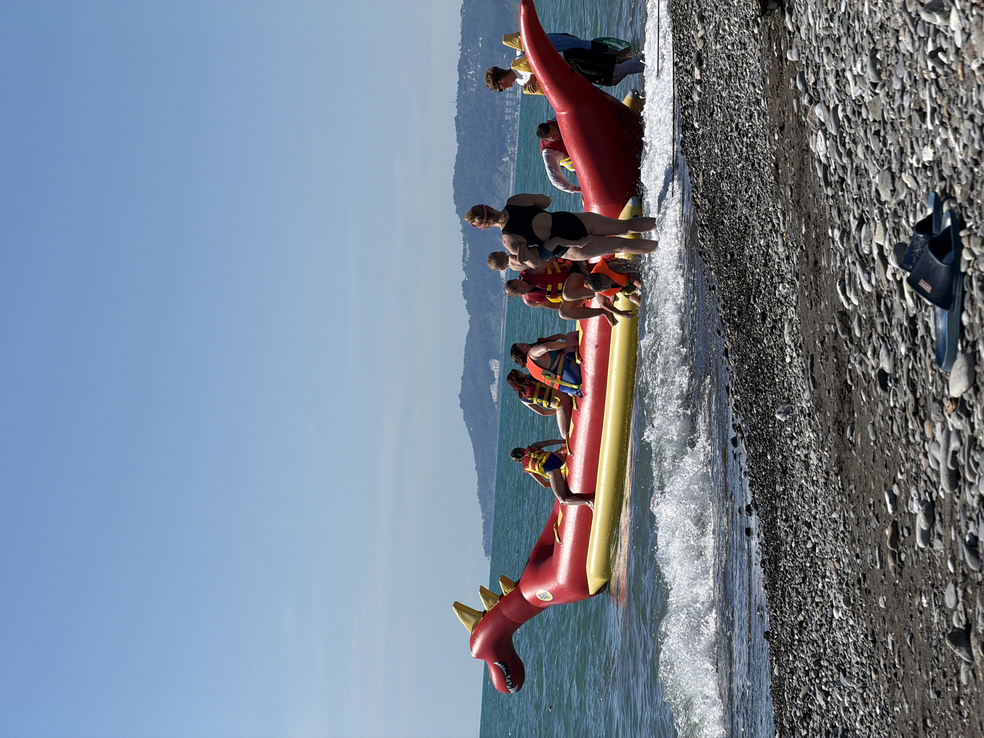 This media file shows a bright, sunny day at a pebbly beach in Sochi, Russia. A group of people are on an inflatable banana boat shaped like a red dragon, which is partially submerged in the blue ocean water. Some of the people are wearing life vests. The banana boat is being pulled by a boat that is out of frame. The water is choppy, with waves crashing onto the shore. The beach is covered in smooth, grey and black pebbles. The sky is clear and blue. In the background, there are hills covered in green trees. One person on the banana boat is holding a child. The overall mood seems to be one of fun and recreation. There is no visible text in the image. The time of day appears to be midday, given the bright sunlight and clear sky.
