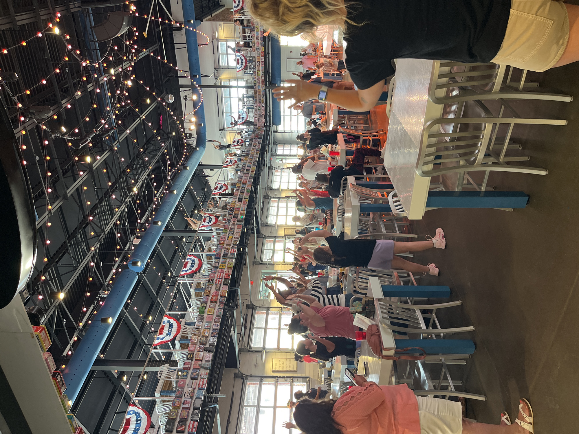This media file captures an indoor gathering at a large, casual venue, likely a restaurant or entertainment space, in Myrtle Beach, USA. The image is taken from an elevated perspective, looking across a room filled with people and festive decor.
**Subjects:** Numerous people of various ages, predominantly adults, are visible throughout the space. Many are standing or sitting at long tables and are engaged in a synchronized activity. In the foreground, the backs of individuals are seen, including a person with blonde hair in a black top and a woman in a pink top using a phone.
**Scene and Setting:** The venue features a high, industrial-style ceiling with exposed dark beams, blue ventilation ducts, and a crisscrossing network of warm-toned string lights, creating a festive and inviting atmosphere. Large windows or glass garage-style doors along one wall allow natural light into the space. The furnishings consist of light-colored tables with blue supports and simple metal chairs. Decorative elements include long displays of small, colorful tins or lunchboxes along a high shelf or beam, interspersed with red, white, and blue patriotic fan decorations.
**Activity:** The central activity involves many people raising their arms above their heads in unison, some with palms together or hands open. This suggests a guided group activity such as a toast, a call-and-response game, a simple exercise, or a celebratory cheer. The atmosphere appears lively and interactive.
**Notable Details:** The abundant string lights are a prominent feature, indicating an evening event or a space designed for a festive mood. The patriotic decorations and unique collection of tins add character to the American-themed setting. On a wall on the right side, a framed sign with text, difficult to read but appearing to be an inspirational message, is visible. An Apple Watch is noticeable on the wrist of one person in the foreground.