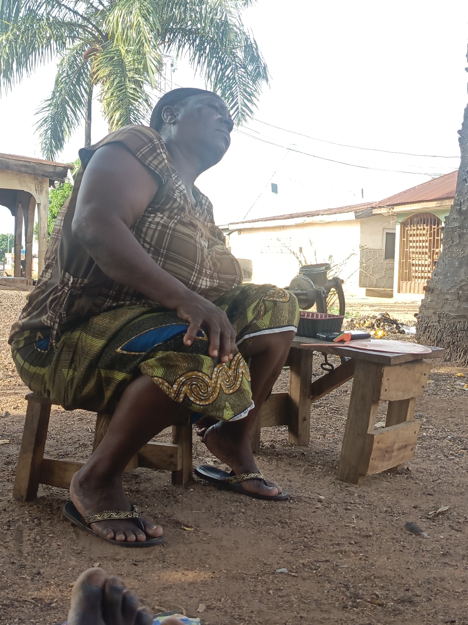 An older, dark-skinned woman is seated on a wooden stool in Akwanga, Nigeria. She is wearing a brown plaid top and a colorful patterned skirt. Her feet are adorned with sandals. Next to her is a small wooden table upon which sits a hand-cranked machine, a container, a calculator, and a small red object. Behind the table is a pile of debris. In the background, there is a building with a barred gate and a white structure. A palm tree is visible overhead. The ground is dirt. The angle of the view is low, with a foot visible at the very bottom.