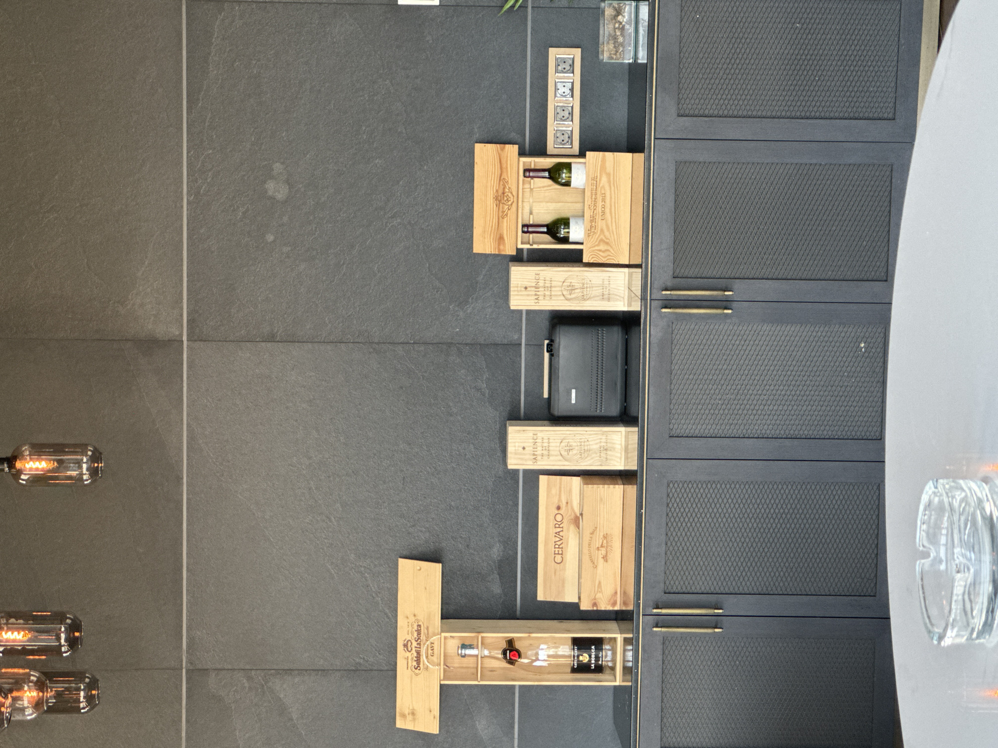 This is a full-shot, eye-level, outdoor photograph taken in Sofia, Bulgaria. The scene depicts a dark grey tiled wall with a horizontal white line in the middle. On a dark grey cabinet with gold handles, there are several wooden boxes with wine bottles. From left to right, there is a wooden box containing a bottle of Gavi wine. Next to it are two stacked wooden boxes with the label "CERVARO." To the right of those are two wooden boxes, one standing upright with the label "SAPIENCE," and another open box containing two green bottles of wine. To the far right, there is a small electrical outlet with four sockets and a small glass terrarium. In the foreground, a round, clear glass ashtray sits on a light-colored circular table. Above the wall, three filament light bulbs are hanging from the ceiling.