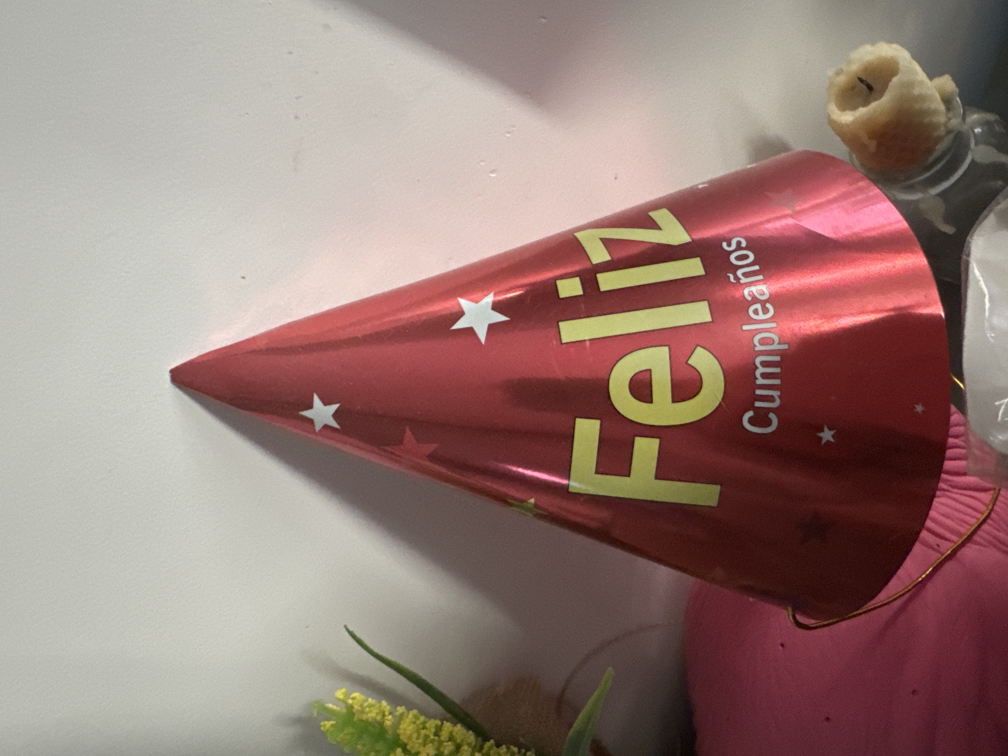 A red, conical party hat with white stars and yellow lettering reading "Feliz Cumpleaños" is placed on a pink surface. The hat is tilted to the right. To the left of the hat, a decorative plant with yellow flowers and green leaves is visible. In the lower right corner, a partially burnt candle is sitting in a clear glass bottle. The background is a plain white wall. The scene appears to be indoors, possibly in a residential setting. There is no information available to place this image in the city of Murcia, Spain.