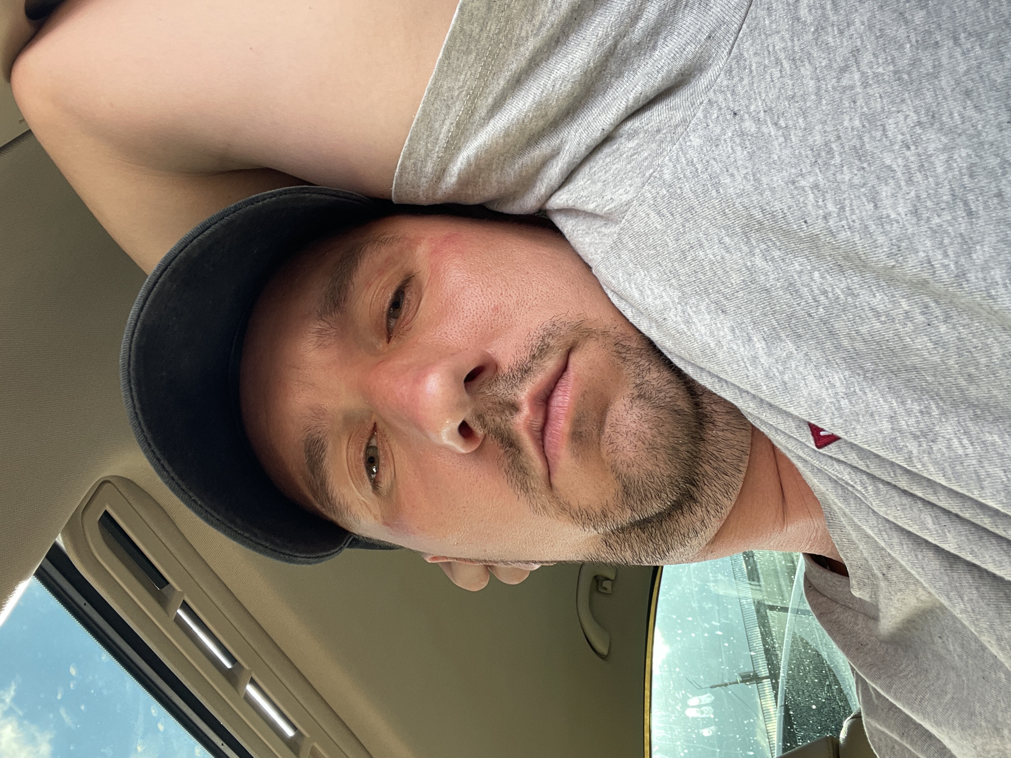 This media file is a close-up selfie of a man wearing a grey t-shirt and a dark baseball cap. He is in a car, as indicated by the car's interior visible in the background, including a sunroof and part of a seat. The man has a short beard and appears to be looking directly at the camera. His facial expression is neutral. The lighting suggests it is daytime, possibly sunny, given the bright sky visible through the car windows. There are some smudges or dirt marks on the car window. The overall mood is casual. The specific location is Kyiv, Ukraine, though this context is not visually evident in the image itself. There is no discernible text in the image.