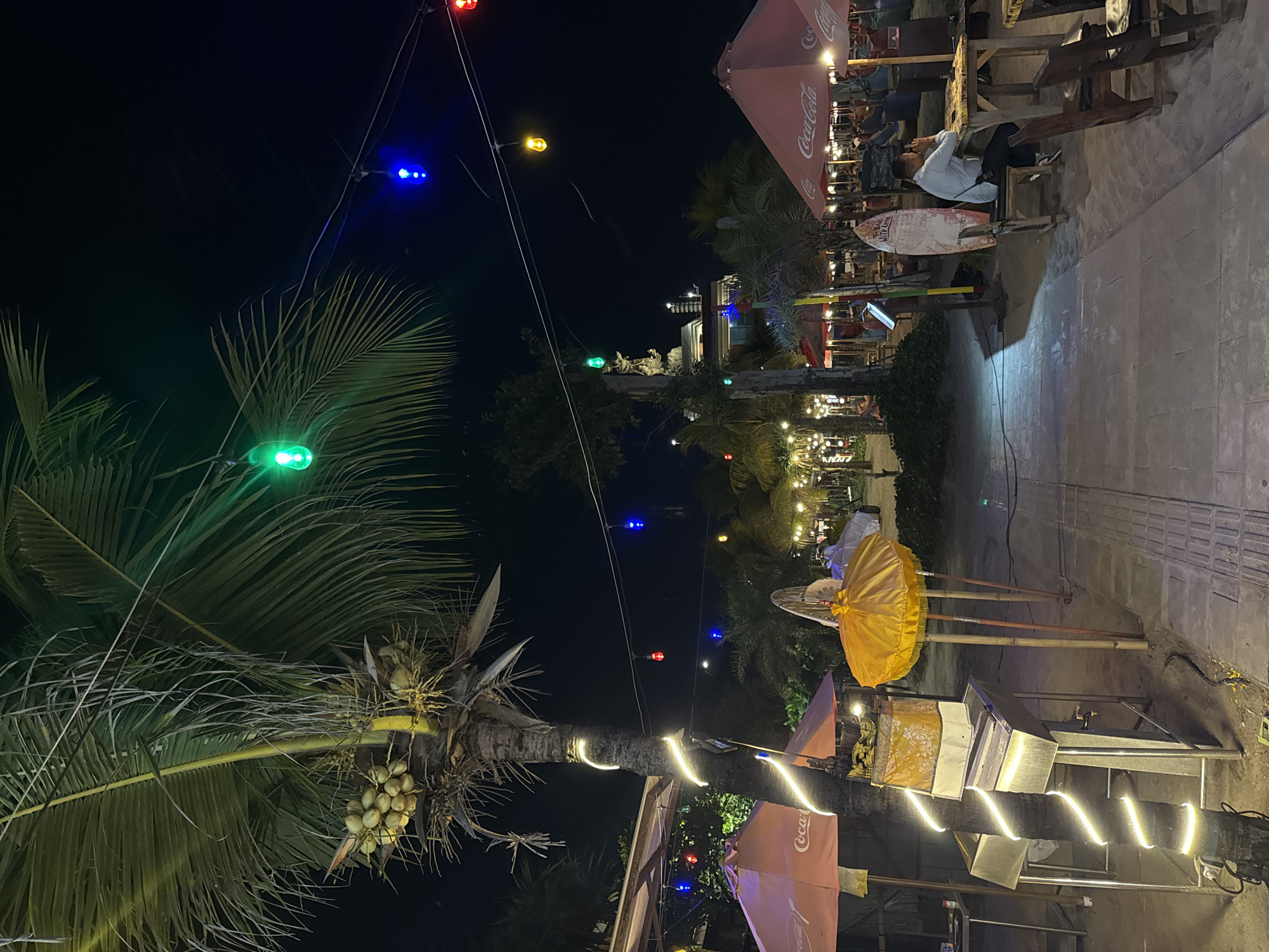 At night, on a beachside promenade in Kuta, Indonesia, a variety of elements create a festive and illuminated atmosphere. In the foreground, a palm tree is wrapped with white string lights, its coconuts visible. To the left, a pink umbrella with "Coca-Cola" branding partially covers a stainless steel food preparation station. Adjacent to this, a bright yellow umbrella is propped up on bamboo poles. Further down the path, several tables and chairs are set up on the sand, some occupied by diners. A surfboard leans against a table where a man in a light blue shirt is seated, looking at his phone. Red, blue, and green string lights are strung overhead, casting a colorful glow on the scene. In the background, other illuminated stalls and structures suggest a lively beachfront establishment. The ground is a mix of paved walkway and sand.