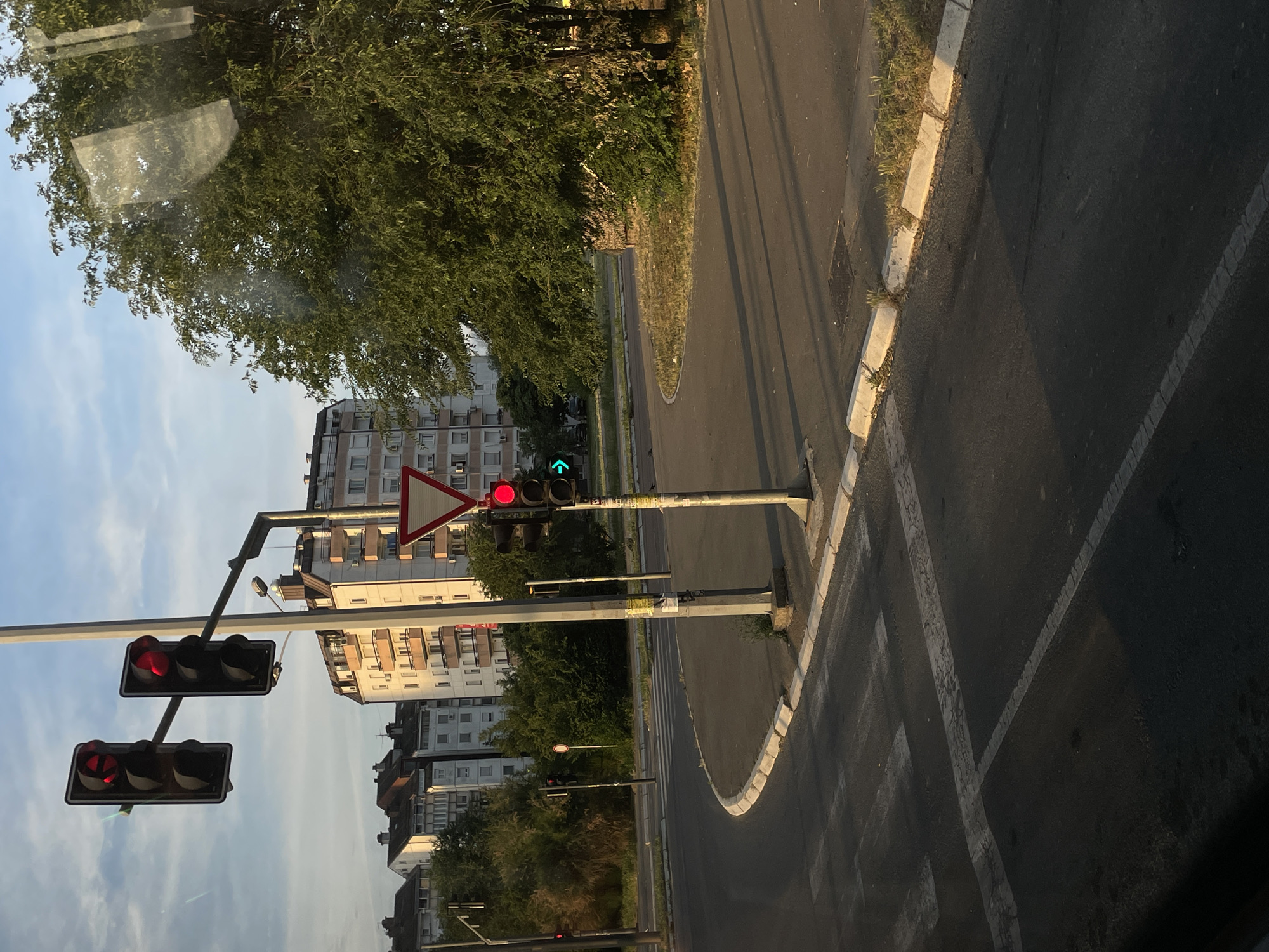 The image shows a street scene in Belgrade, Serbia. Two traffic lights are visible, one showing red lights and the other showing a green arrow, indicating a turn. A yield sign is also present. Buildings are visible in the background, suggesting a residential or mixed-use area. The road surface is asphalt, and there is a sidewalk with a curb. Trees and vegetation are also part of the environment. The photo seems to have been taken from inside a vehicle.