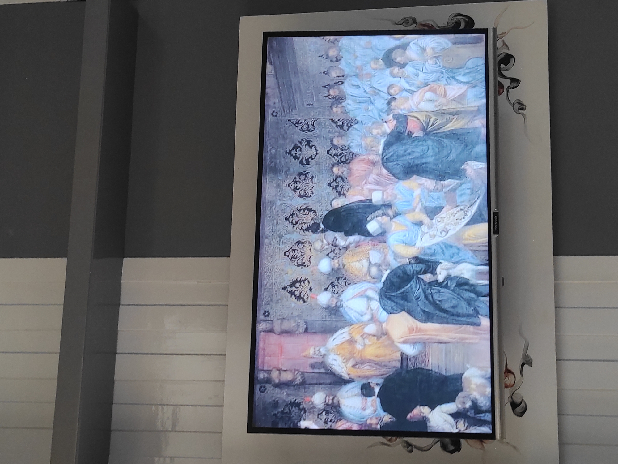 The image shows a television screen displaying a painting of a historical scene with multiple figures in elaborate, traditional attire. The painting appears to depict a gathering or ceremony, with a central figure in a golden robe and ornate headwear, possibly royalty. Other figures in the painting are dressed in various colored robes and head coverings, some in shades of blue, orange, and white. The background of the painting features intricate patterns and architectural elements. The television itself is mounted on a wall decorated with a modern, abstract design in gray and white. The wall beneath the television has vertical white panels. The overall setting appears to be a contemporary interior space. The painting is rich in detail, suggesting a narrative or ceremonial event. There are also two dogs visible in the foreground of the painting. There is no indication that this scene is located in Sumedang, Indonesia.