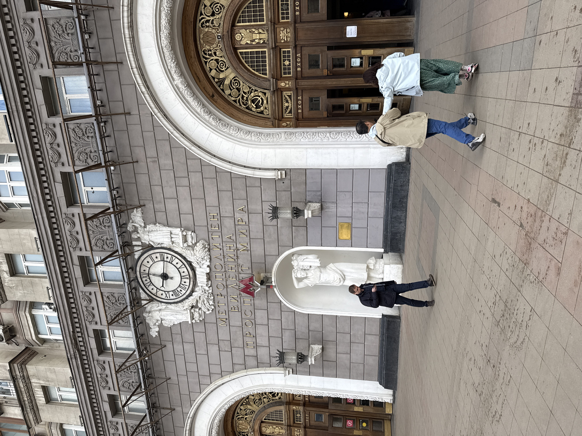 The image shows the entrance to the Prospekt Mira Metro station in Moscow, Russia. The building is a grand, grey stone structure with ornate architectural details, including arched entryways, sculptures, and a large clock above the central entrance. A sign in Cyrillic above the main entrance identifies the station. Two people are entering the right-side doorway and one person is walking away from the central entrance. The exterior pavement is made of square tiles.