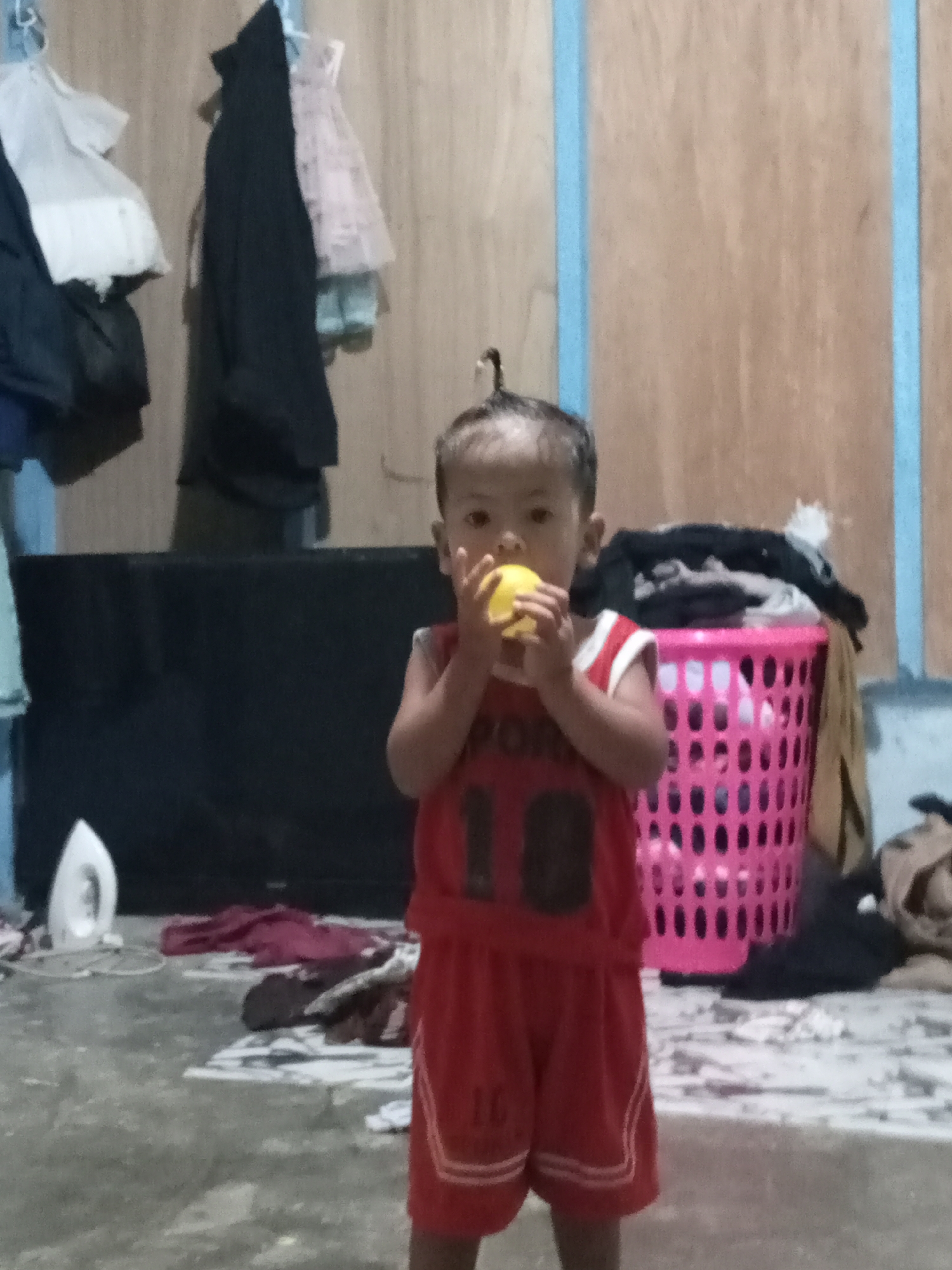 A young child, wearing a red athletic outfit, stands in the center of the frame holding a yellow ball up to their mouth. The child's hair is pulled up into a small bun. Behind the child, a pink plastic laundry basket is filled with clothes. To the left of the child, a white iron is visible on the floor. The background consists of a wooden wall with blue accents and several garments hanging on hooks. The floor is concrete and appears somewhat cluttered with fabric. The overall setting suggests a domestic, likely indoor, environment.