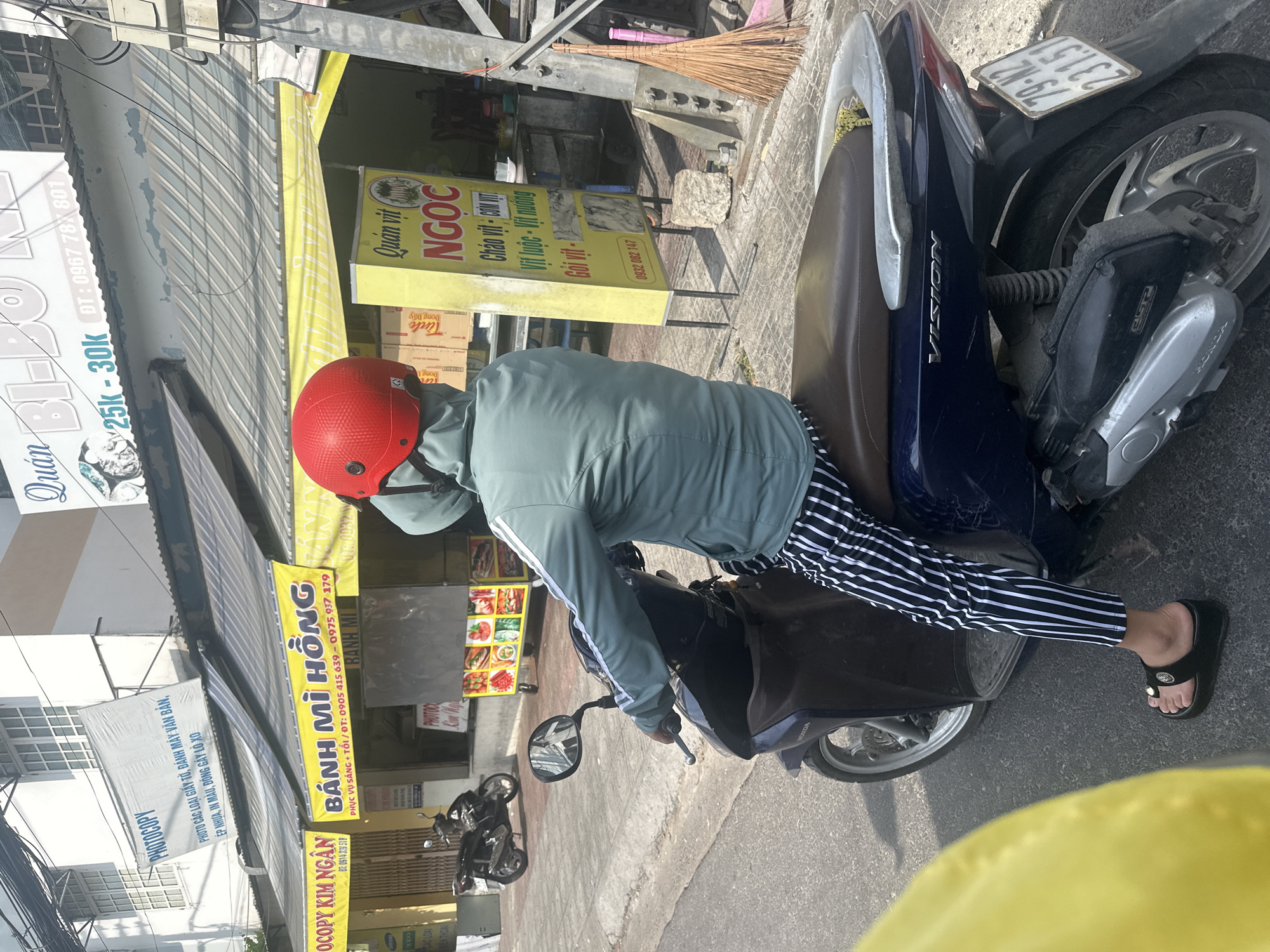 A person wearing a teal hooded jacket, black and white striped pants, and black platform sandals is seated on a dark blue Honda Vision scooter. The scooter has a license plate reading "79-N2 231.51". The scooter is parked on a paved street in front of a row of shops. A yellow banner with Vietnamese text and red lettering reads "BÁNH MÌ HỒNG". Another yellow sign, also with Vietnamese text, displays "Quán vịt NGOC". In the background, there is a building with multiple windows and a blue awning. A broom made of twigs leans against a metal pole to the right of the frame. The scene appears to be a street in Bac Nha Trang Ward, Vietnam.