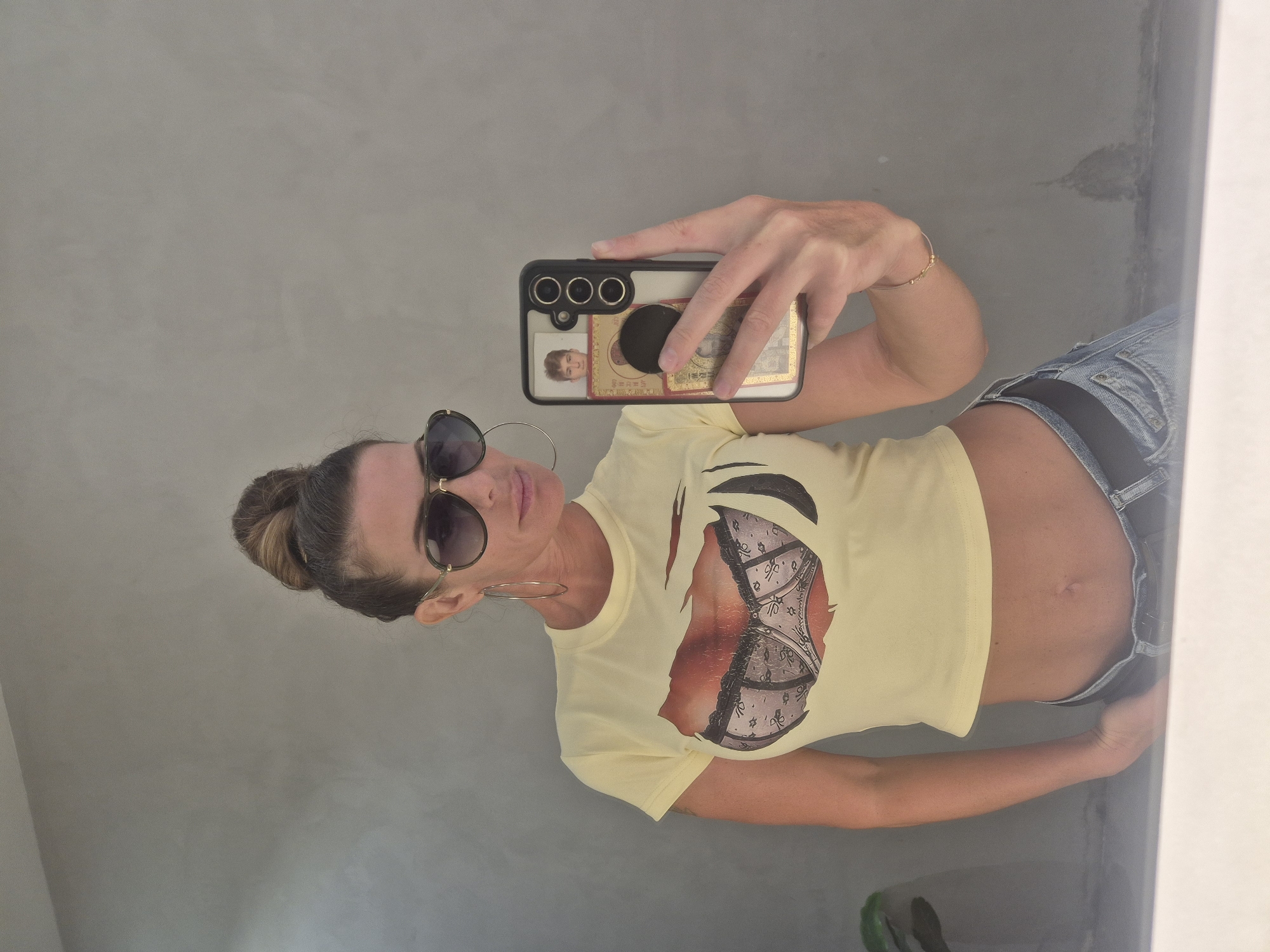 A woman is captured in a mirror selfie, appearing horizontally oriented in the image. She has light skin, hair pulled back into a bun with highlights, and is wearing large, dark-tinted sunglasses and silver-colored hoop earrings. Her lips are colored a light pink. She wears a light yellow crop top featuring a graphic of a ripped shirt exposing a lacy bra underneath. Her midriff and navel are visible. She is also dressed in light blue denim shorts and has a simple gold-colored bracelet on her right wrist.
She holds a smartphone in her left hand, which has a clear case. Inside the case, a small photo of a person (likely a young individual with light hair) and a golden rectangular item with faint, indiscernible text and patterns are visible, along with a black phone grip. The background is a plain, light grey or beige wall, suggesting an indoor setting. The location context is Dubai, United Arab Emirates. The activity depicted is a casual self-portrait.