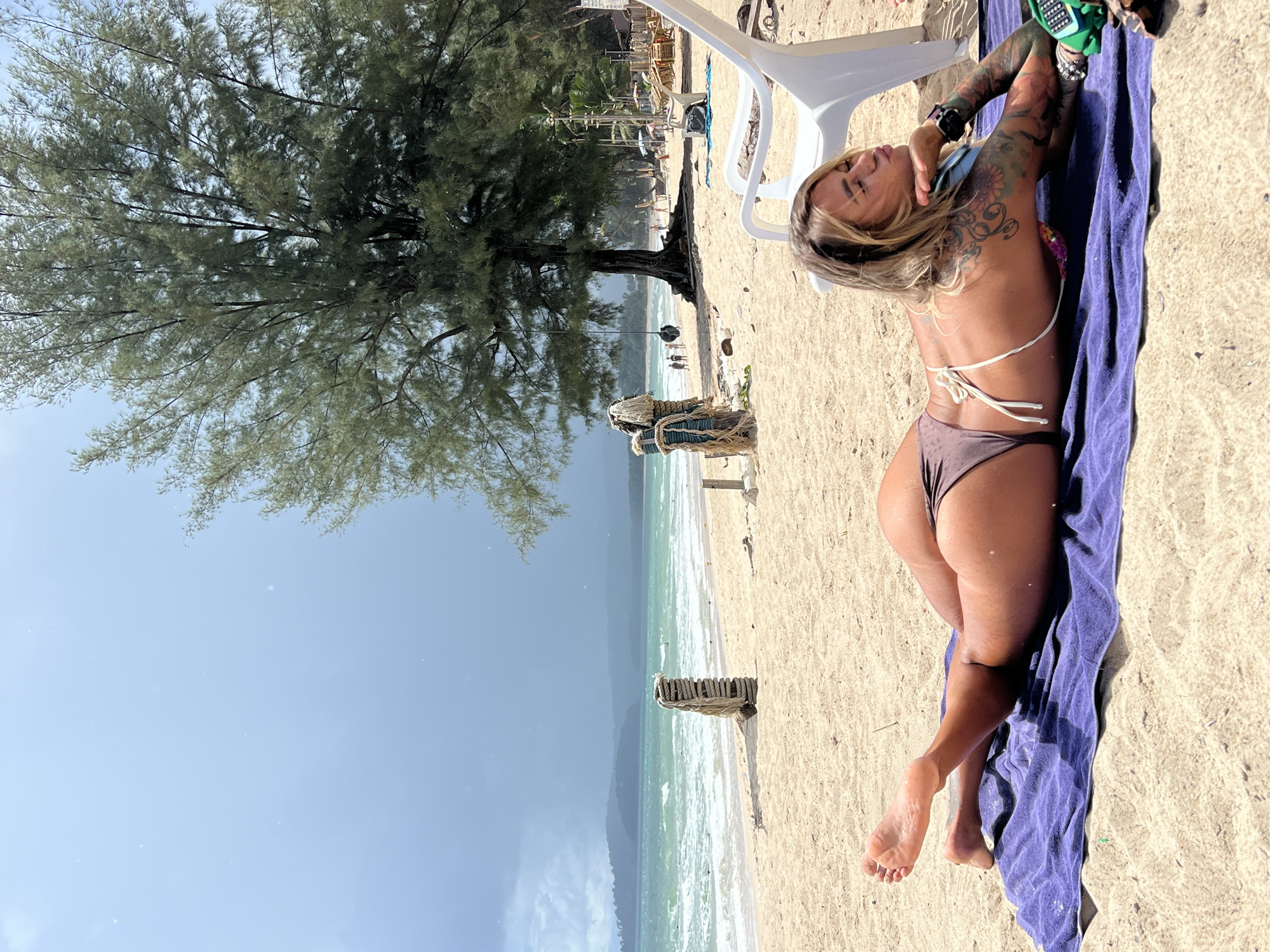 An outdoor, eye-level, medium shot shows a woman lying face down on a purple towel on a sandy beach. The woman is wearing a brown bikini. She is positioned in the lower right quadrant of the frame and is looking towards the right. Her head is turned to the right, and her left arm is bent, with her hand resting on her chin. Her right arm is extended forward, with her hand resting on the towel. She has blonde hair and tattoos on her right arm. The beach is light brown sand and there is the ocean in the background with light turquoise water and white waves. There are several trees and a white plastic chair visible. The sky is light blue with some grey clouds in the distance.