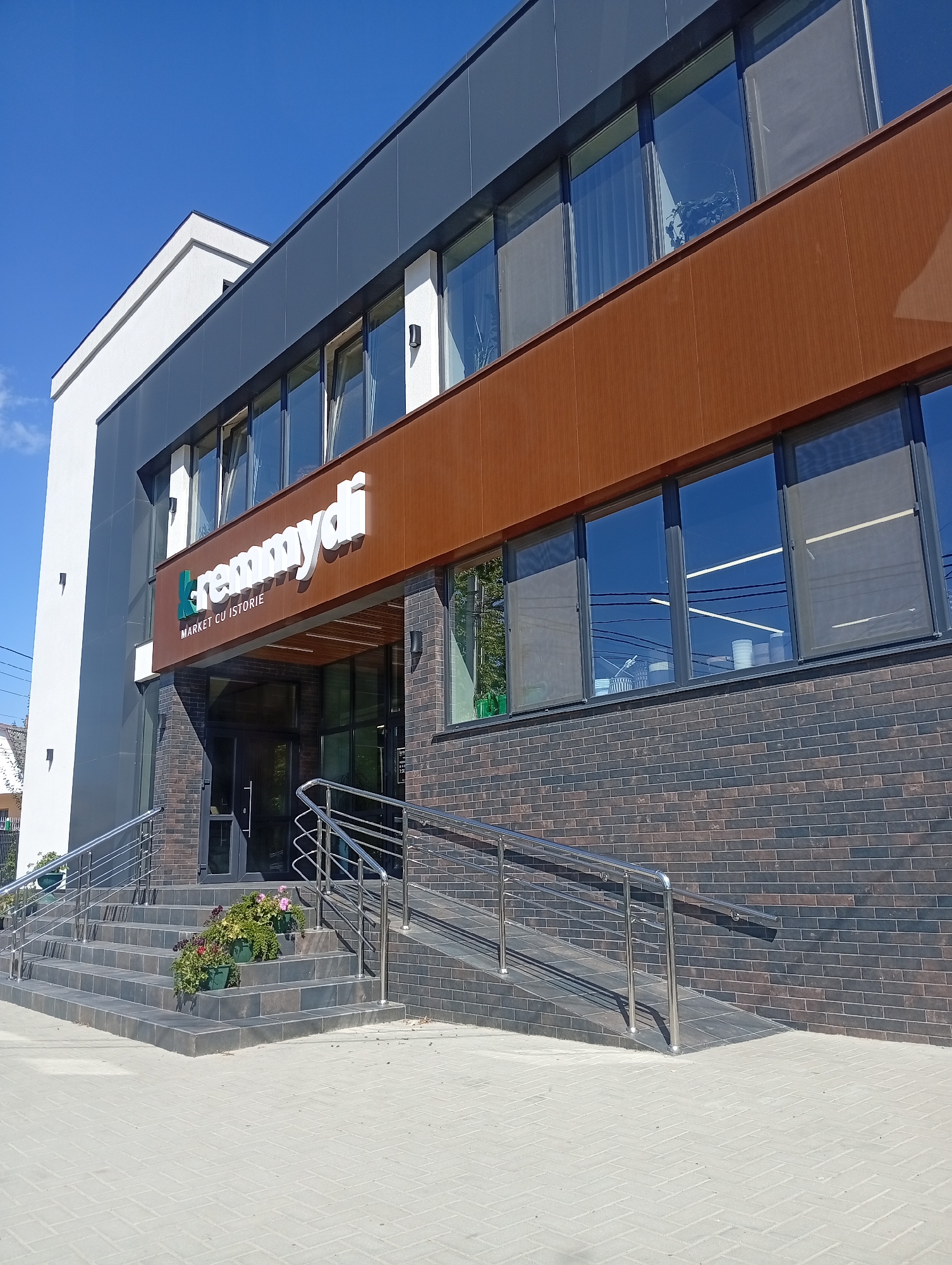 The image shows a modern building exterior with a sign that reads "remmydi MARKET CU ISTORIE". The building features a dark gray facade, a brown horizontal accent panel, and large windows. To the left of the entrance, there are stairs leading up to the building, flanked by chrome railings. Potted plants with pink and red flowers are placed on the stairs. A ramp with chrome railings is visible to the right of the stairs. The ground in front of the building is paved with light gray interlocking bricks. The sky is clear and blue. The building is located in Pănășești, Moldova.