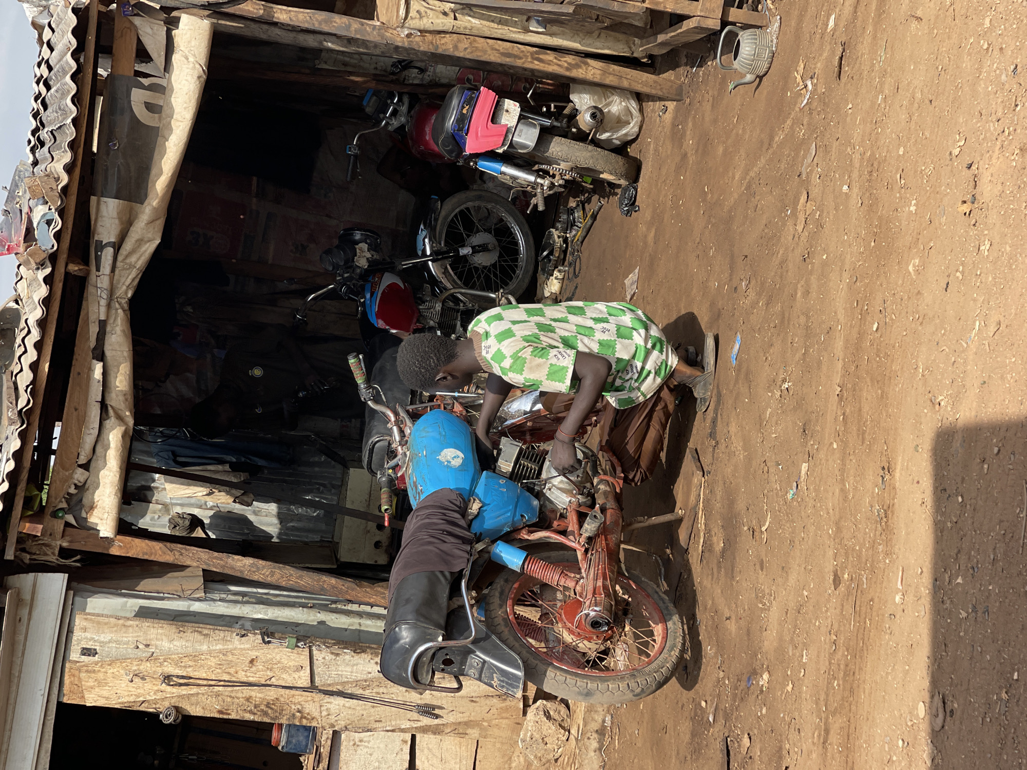 A young person wearing a green and white checkered shirt kneels while working on a red and blue motorcycle in a makeshift workshop. The workshop is constructed with corrugated metal and wood. Several other motorcycles are parked behind the one being repaired. The ground is dirt and gravel. The location appears to be in Abuja, Nigeria.