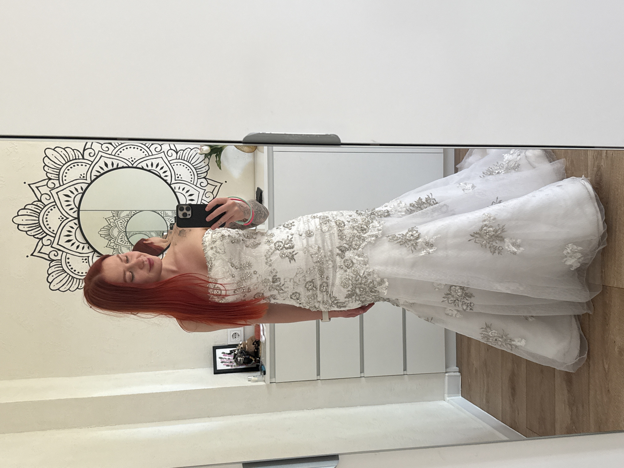A red-haired female stands in a domestic indoor setting located in Rostov-on-Don, Russia, taking a mirror selfie. She has long, straight auburn hair, fair skin, and a stud piercing on her right nostril. Tattoos are visible on her left upper arm/shoulder and covering a significant portion of her left forearm, along with several colorful bracelets (neon green, pink, orange) and a white smartwatch on her left wrist. She is wearing a strapless white wedding gown featuring intricate silver and white floral embroidery, beading, and sequin embellishments, designed in a mermaid or trumpet silhouette with a flared skirt. She holds a black smartphone with a triple-lens camera in her left hand, angled towards her reflection in a large, clear mirror. Her eyes are downcast, and she exhibits a slight, closed-mouth smile.
The environment behind her reflection includes a white wall adorned with a large black and white mandala-style decal centered around a smaller circular mirror. To her left, a white chest of drawers with four visible drawers holds a black-framed picture displaying abstract pink and purple elements, several small containers, and a white electrical outlet. To her right, a single white tulip in a gold-colored vase rests on a white surface. The floor is composed of light brown, wide plank wood. A plain white door panel with a vertical dark gray handle is visible on the far right, suggesting the main mirror is part of a wardrobe or closet door.