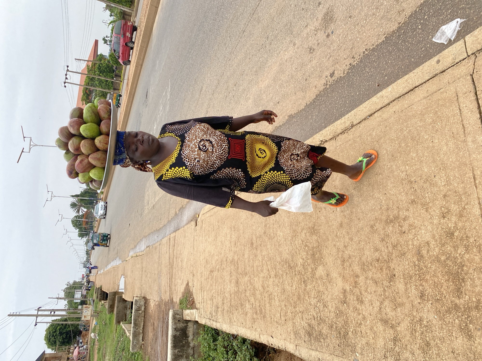 A medium shot portrays a woman carrying a large tray of round, green and red fruits on her head. She is walking along a concrete sidewalk in Bukuru, Nigeria. The woman is wearing a dark dress with a pattern of brown and yellow circles, and orange flip-flops. She holds a white plastic bag in her left hand. The sidewalk is next to a street with cars and a green Tuk-tuk (auto rickshaw) in the distance. Utility poles line the street. The sky is overcast.