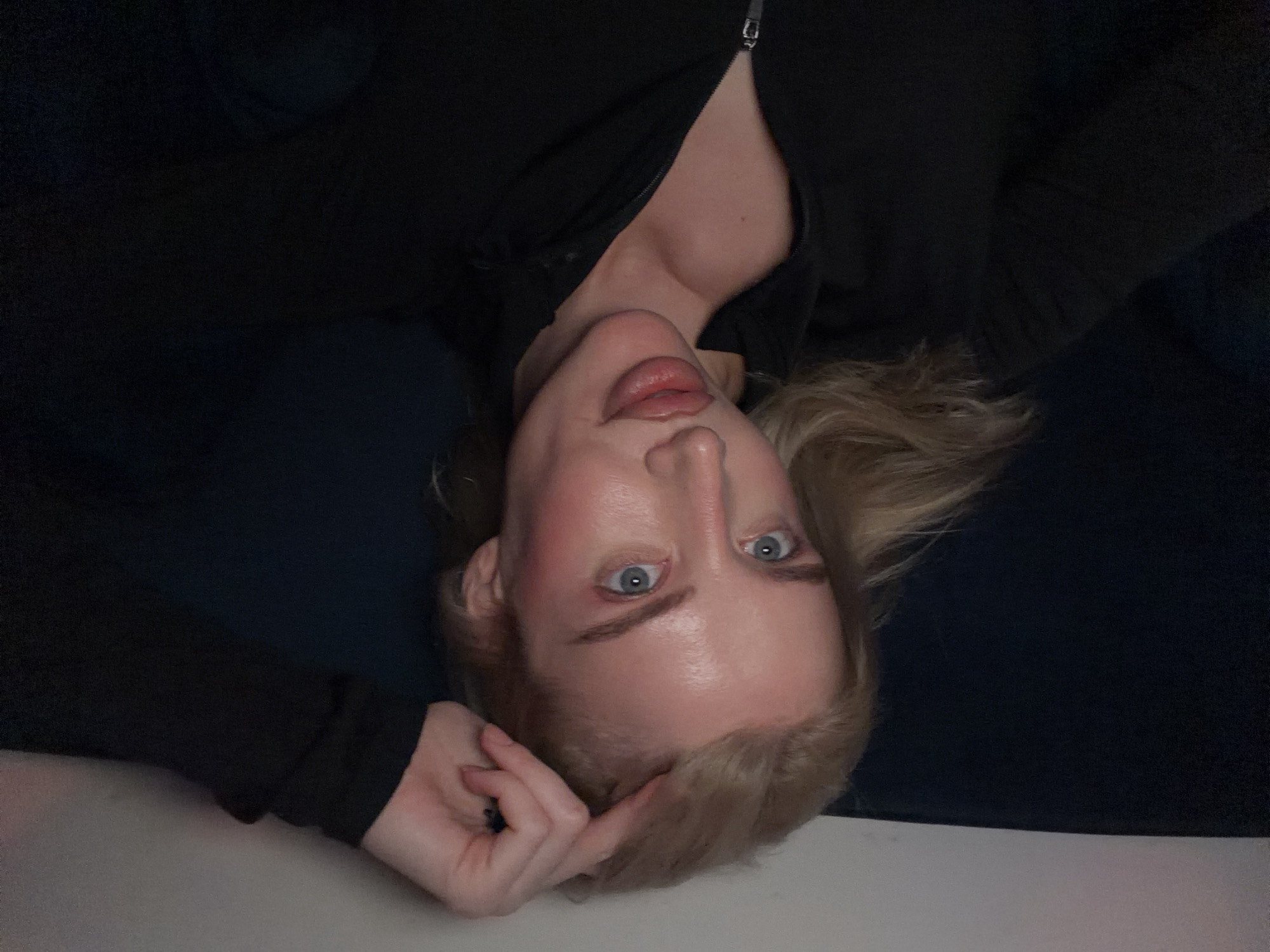 The image displays a person, likely female, with fair skin, light blonde hair, and wide blue eyes, lying on her back and looking directly into the camera. She is positioned upside down within the frame, with her head near the bottom edge. She is wearing a dark, possibly black, long-sleeved top or jacket, featuring a visible silver zipper near her neck and chest area. Her lips are slightly parted, and she appears to have light makeup, including blush on her cheeks and lip color. Her right hand is gently resting near her head, touching her hair. The background is predominantly dark, suggesting an indoor setting with low or ambient lighting. A lighter surface, presumably where she is lying, is visible along the bottom edge of the image. The activity is a posed photograph, possibly a selfie. There are no specific architectural or geographical cues within the image to indicate Belgrade, Serbia. No text is visible in the image.
