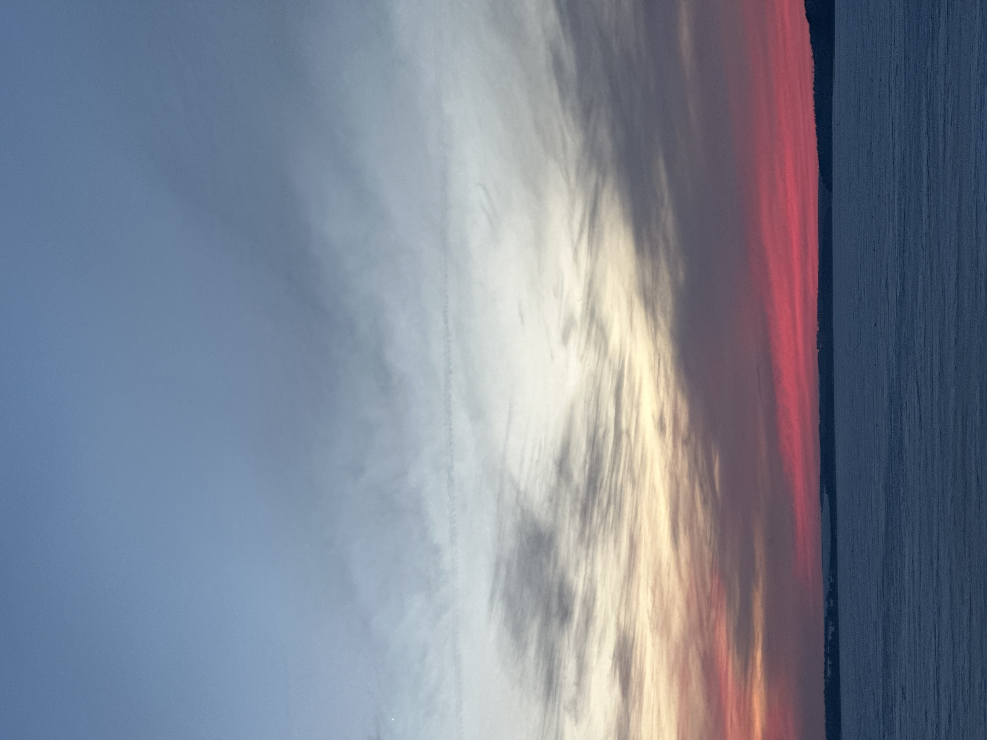 The image displays a wide, static, landscape view of a frozen body of water under a colorful sky, located in Votkinsk, Russia. The sky, occupying the upper two-thirds of the frame, exhibits a gradient from deep blue at the top, transitioning through pale blue-grey and white with wispy cirrus clouds, to distinct horizontal bands of pale yellow, orange, and intense red above the horizon. A faint, linear contrail is visible horizontally across the upper-middle sky. The horizon line reveals a dark, low-profile landmass, likely a distant treeline, stretching across the entire width of the scene. The foreground, comprising the lower third of the image, consists of a vast expanse of uniform dark blue-grey frozen water or snow-covered ice, showing subtle surface texture from parallel lines or ridges. No individuals, man-made structures, or distinct actions are depicted.