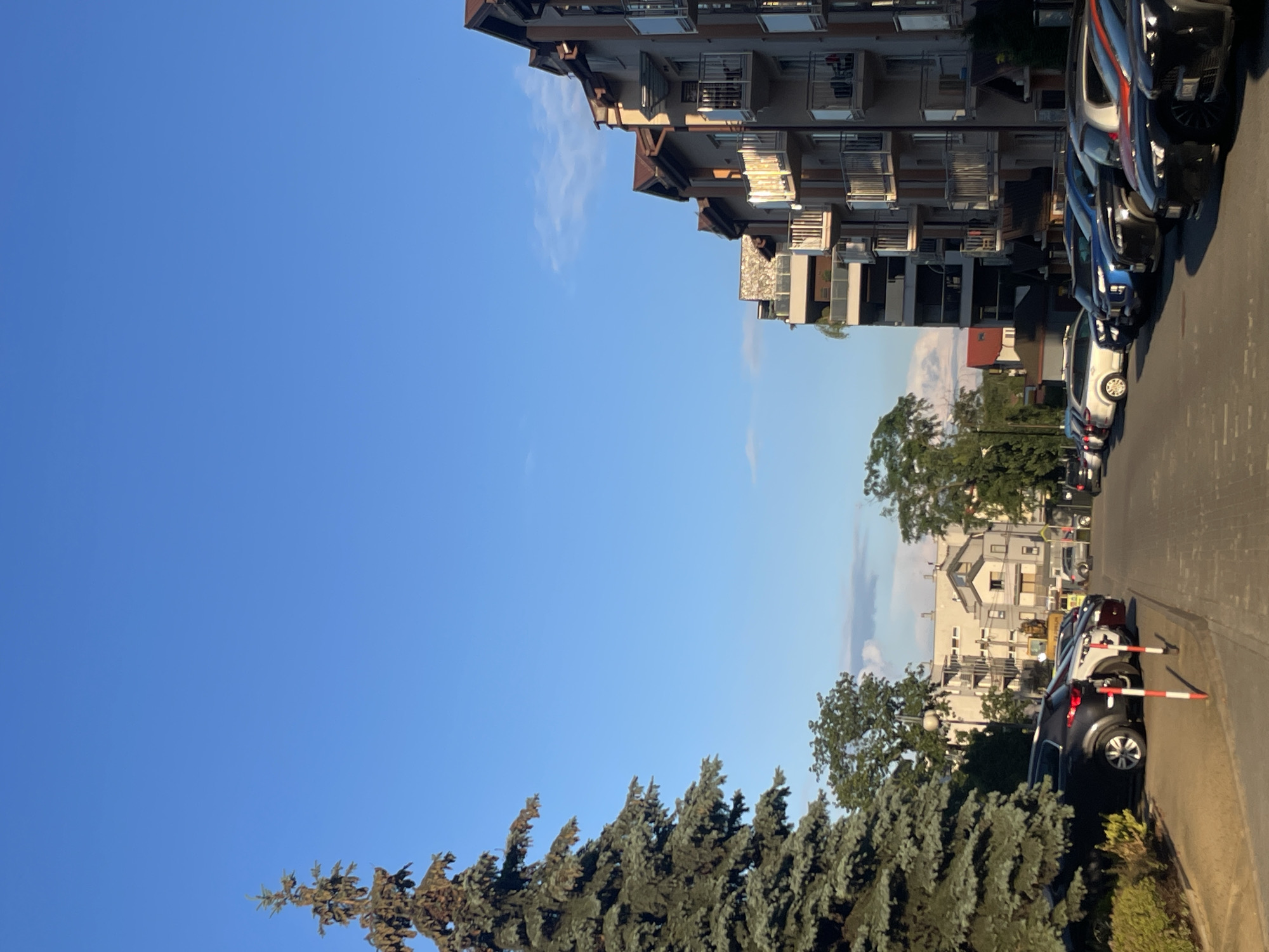The image depicts an outdoor residential scene under a clear, bright blue sky with minimal scattered white clouds. A large, mature coniferous tree with dark green needles occupies the left side of the frame, extending from the bottom to near the top. On the right, a series of modern multi-story apartment buildings with light-colored facades, prominent brown pitched roofs, and multiple balconies featuring glass and metal railings are visible. Direct sunlight illuminates parts of these buildings, particularly the balconies.
The central and foreground areas show a paved street or parking lot densely populated with parked passenger vehicles, including SUVs and sedans, in various colors such as black, white, blue, and red. These vehicles are arranged along both sides of the thoroughfare. Red and white striped bollards are present on the left side of the road, near the curb and the base of the large tree. In the mid-ground, beyond the parked cars and immediate buildings, further residential structures, smaller trees, and foliage are visible. A yellow construction vehicle can be seen in the distance, slightly left of the center. No individuals are discernible in the scene. The lighting suggests late afternoon.
Location: Residential area within the city of Unknown, Unknown.
