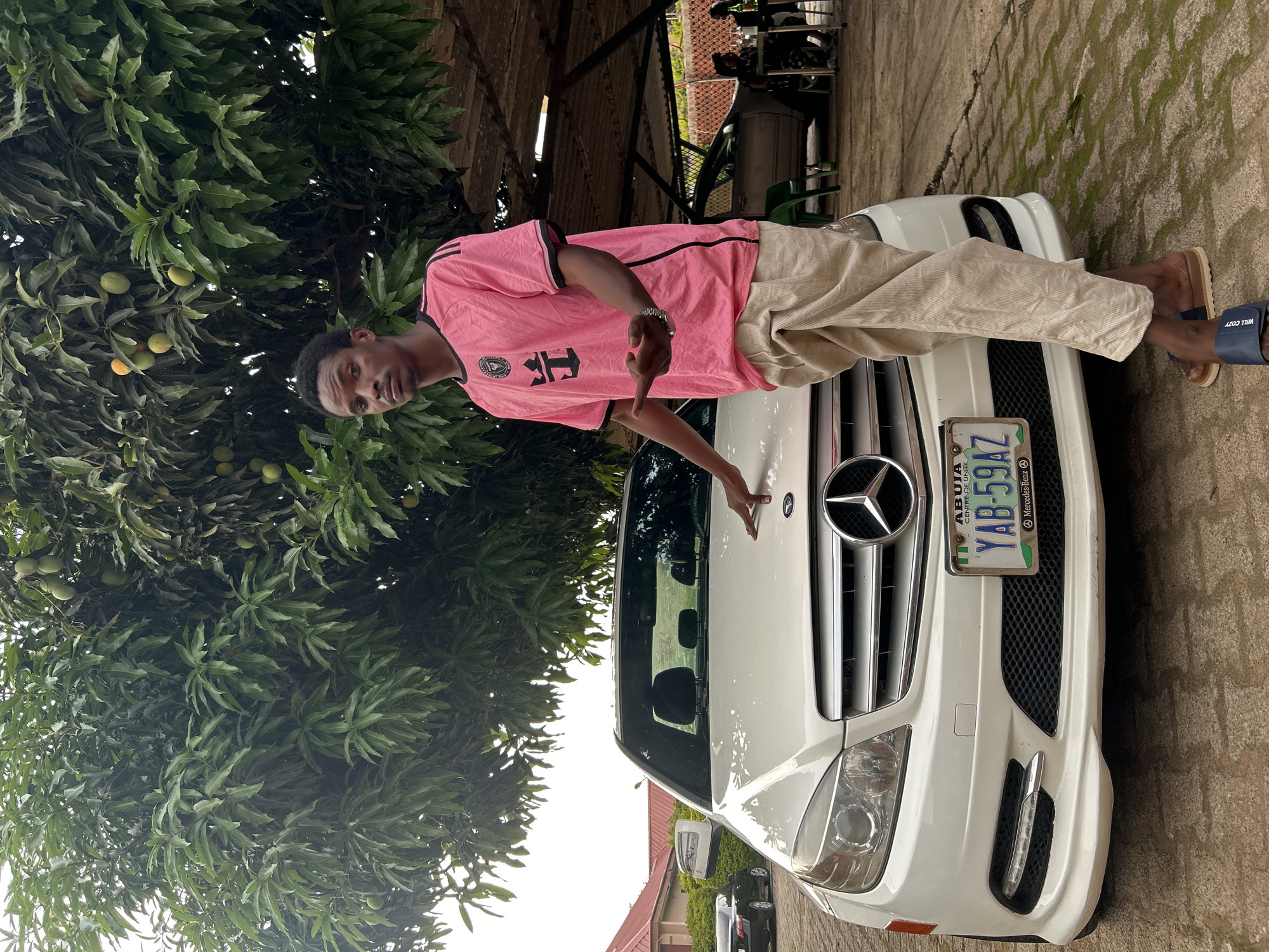 A young man stands next to a white Mercedes-Benz with an Abuja license plate (YAB-594AZ). He wears a pink shirt with black trim and khaki pants, points with both hands towards the car's hood. The car is parked on a paved surface, with a large leafy tree to the left, partially obscuring the background. The location is in Jos, Nigeria.