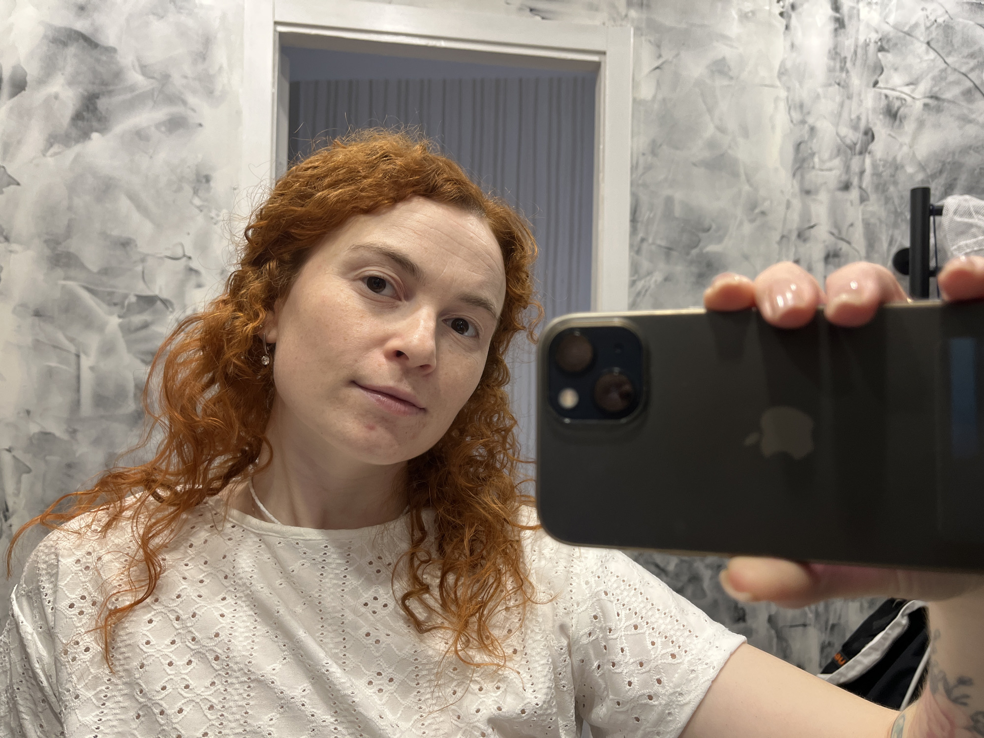 A person with red, curly hair is taking a selfie with a black iPhone. The person is wearing a white, eyelet-style top. The background shows a white doorframe and a wall with a textured, gray and white pattern. The location appears to be indoors, possibly a bathroom. There is no clear evidence to suggest this image was taken in the city of Chaikovsky, Russia.