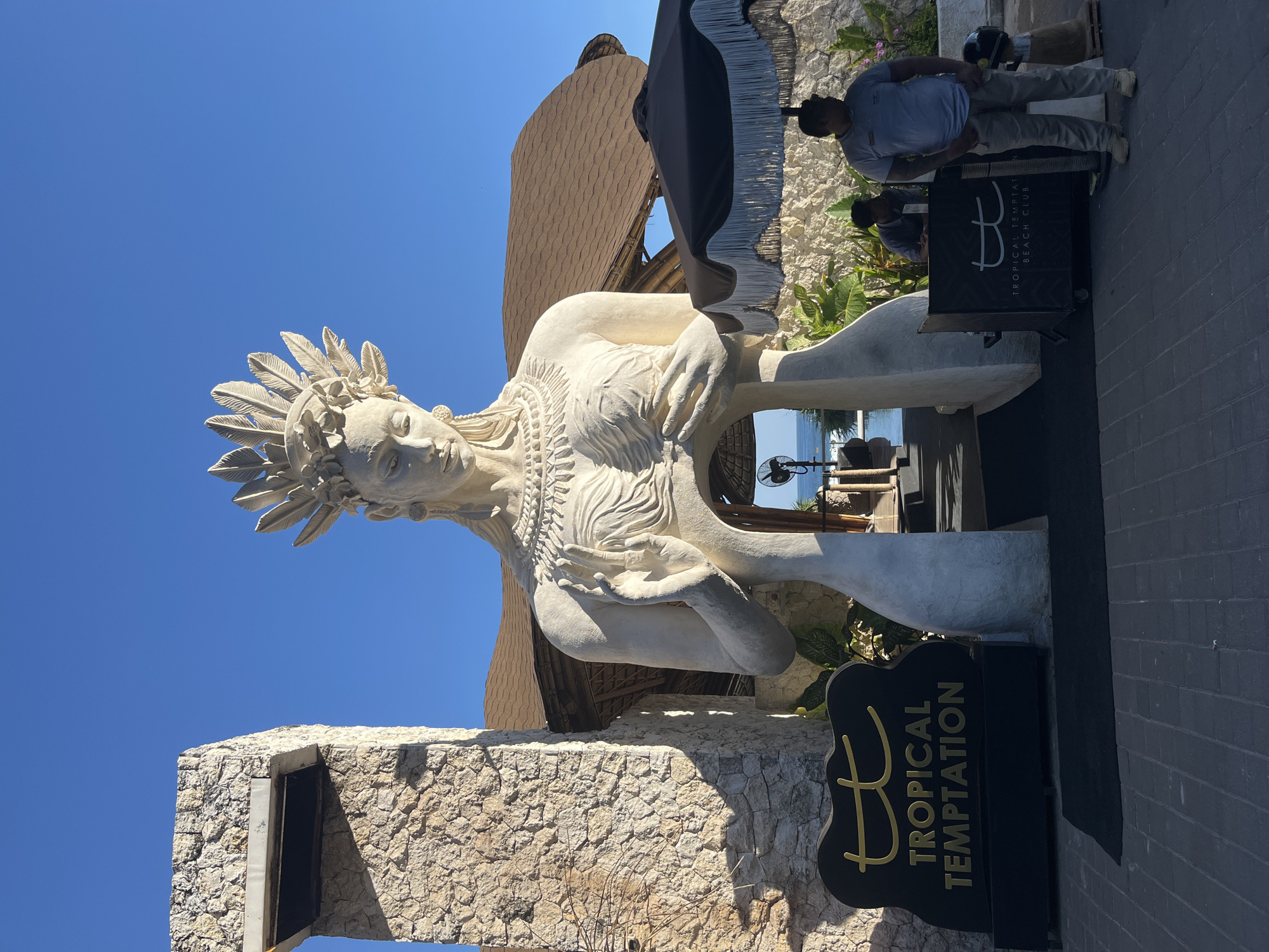 A large, white stone statue of a seated female figure with a feathered headdress is the central subject of this outdoor scene. The statue is intricately carved, depicting detailed facial features, elaborate jewelry around the neck, and the texture of feathers. The figure's hands are crossed over its chest, and its gaze is directed downwards.
The statue is positioned in front of a building with a distinctive curved, thatched roof and walls made of rough-hewn stone. Behind the statue, lush green foliage is visible, suggesting a tropical environment. In the distance, a glimpse of the ocean and a clear blue sky indicate a sunny day.
Two men are partially visible in the background. One man, wearing a light blue t-shirt and grey pants, stands near a black sign. Another man, also in a t-shirt and pants, is seated behind him.
A black sign with gold lettering stands to the right of the statue. The text on the sign reads "TROPICAL TEMPTATION BEACH CLUB". The overall atmosphere appears to be that of a resort or beach club, likely during daytime given the bright sunlight and clear sky. The scene is set in Kuta Selatan, Indonesia.