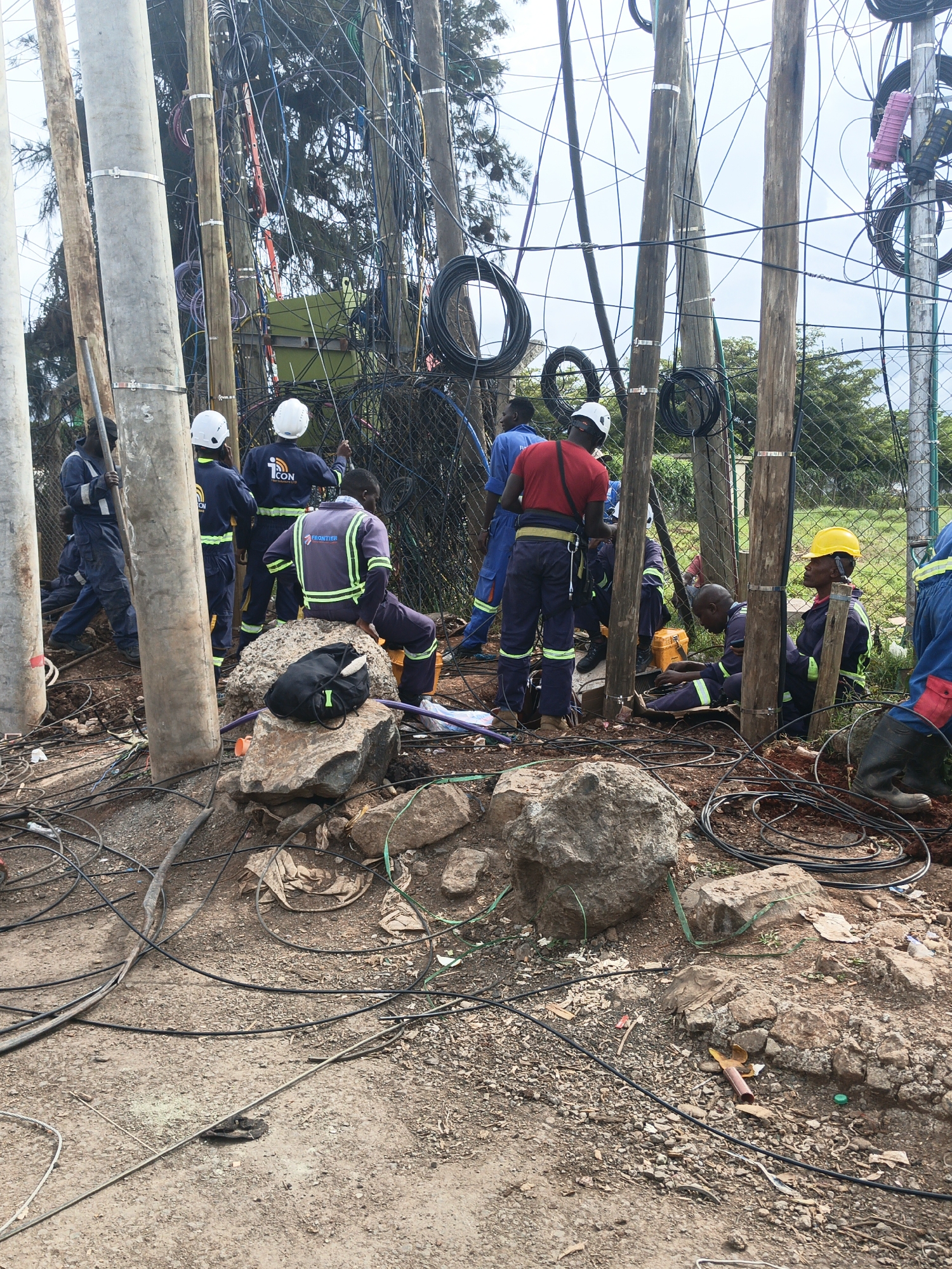 The image captures an outdoor scene in Nairobi, Kenya, where a group of approximately ten to twelve male workers are engaged in what appears to be maintenance or installation work on a complex network of utility poles and cables. The scene is dominated by several tall poles, a mix of concrete and weathered wood, heavily laden with a dense, tangled web of black, blue, and multicolored electrical and communication wires. Numerous coils of black cable are also visible attached to the poles.
The workers are dressed in work uniforms, mostly blue or grey overalls, with some featuring high-visibility reflective stripes. Many wear white hard hats, and one worker is seen in a yellow hard hat. Two distinct company logos are visible: one worker's uniform displays "iCON" with a network globe icon, and another worker's uniform has "Frontier" written on it.
The men are positioned around the base of the poles, with some standing, some crouching, and others seated on large, rough rocks scattered across the dusty, reddish-brown ground. Tools, including a yellow toolbox, are present. In the background, a chain-link fence is visible, with trees and green foliage beyond it. A large green utility box or transformer is mounted high up on one of the wooden poles, almost hidden amidst the wires. The sky is overcast, suggesting a cloudy day. The ground is uneven, covered in dirt, small rocks, and some scattered debris, including loose cable segments. The overall impression is one of intensive manual labor involved in managing intricate infrastructure.