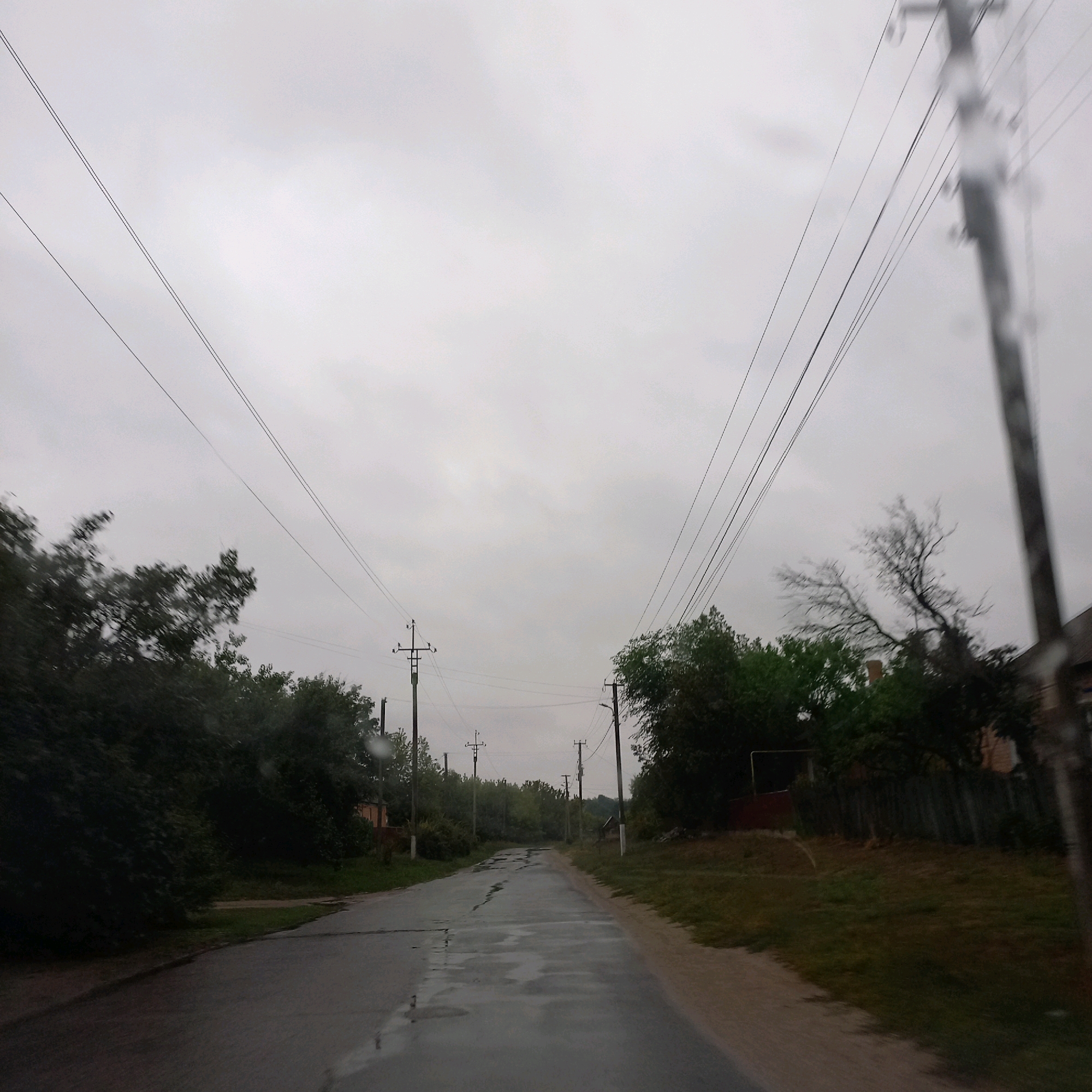 The image depicts a straight, paved road in a rural or suburban setting, likely on a cloudy day with signs of recent rain, indicated by the wet surface and reflections on the asphalt. Power lines are prominent, strung between utility poles that line the road. Trees and vegetation border the road, with some appearing lush and green, while others are bare. A wooden fence is visible on the right side of the road, and a building with a brick facade is partially visible behind it. The overall scene suggests a quiet, possibly residential area. There are no people visible in the image.