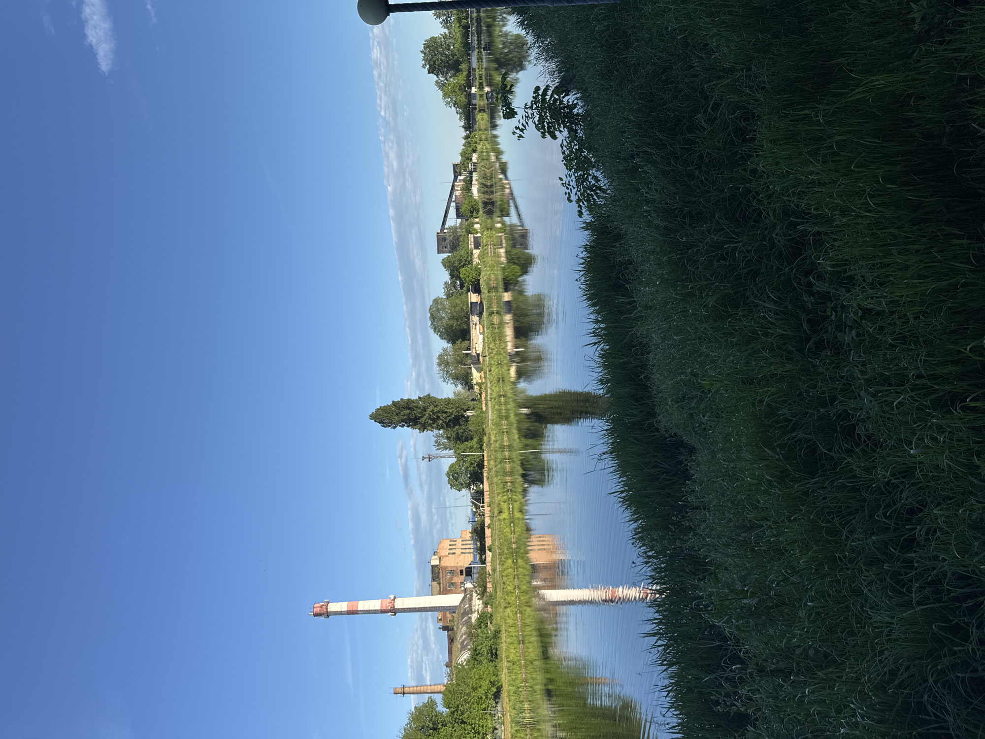 The image shows a body of water with green vegetation along the bank. The water reflects the sky and a building with a tall chimney. The sky is blue with a few clouds. A street lamp is visible at the top. The scene appears to be taken in an urban or industrial area.