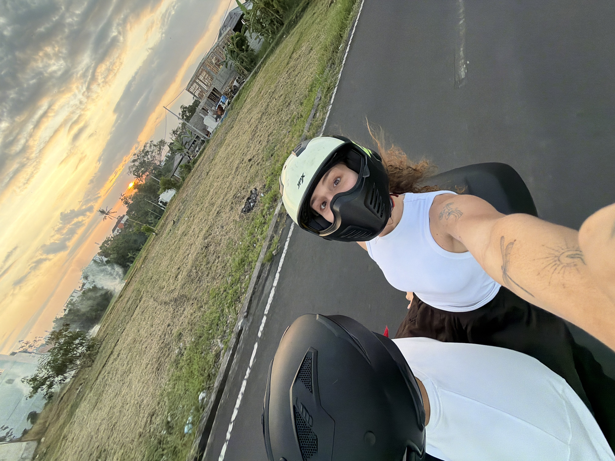 This media file depicts two individuals riding on a motorbike, with a selfie-style perspective. The subject in the foreground, wearing a white tank top and black pants, is looking directly at the camera. They are wearing a light-colored helmet with a black face shield. A tattoo of a dragonfly and a radiating sun is visible on their arm. Behind them, another person wearing a black helmet is partially visible, also in a white top.
The scene is set outdoors during what appears to be either sunrise or sunset, with a dramatic sky filled with elongated clouds illuminated by golden and orange hues. The sun is low on the horizon, casting a warm glow. The motorbike is on a paved road, with a grassy field to the left. The field is somewhat unkempt, with patches of dried grass and some debris. In the background, there are indications of construction, with unfinished buildings visible amongst trees. Further in the distance, some structures and palm trees suggest a tropical or semi-tropical environment. A plume of smoke is visible rising from the grassy area, possibly indicating controlled burning or a small fire.
The overall mood of the image is casual and adventurous, given the motorbike ride and the individuals' attire. The lighting suggests a peaceful, transitional time of day. The presence of unfinished construction alongside natural scenery hints at development in the area. The text "JPX" is visible on the white helmet in the foreground. The location context of North Kuta, Indonesia, aligns with the visual cues of tropical vegetation and the general aesthetic.