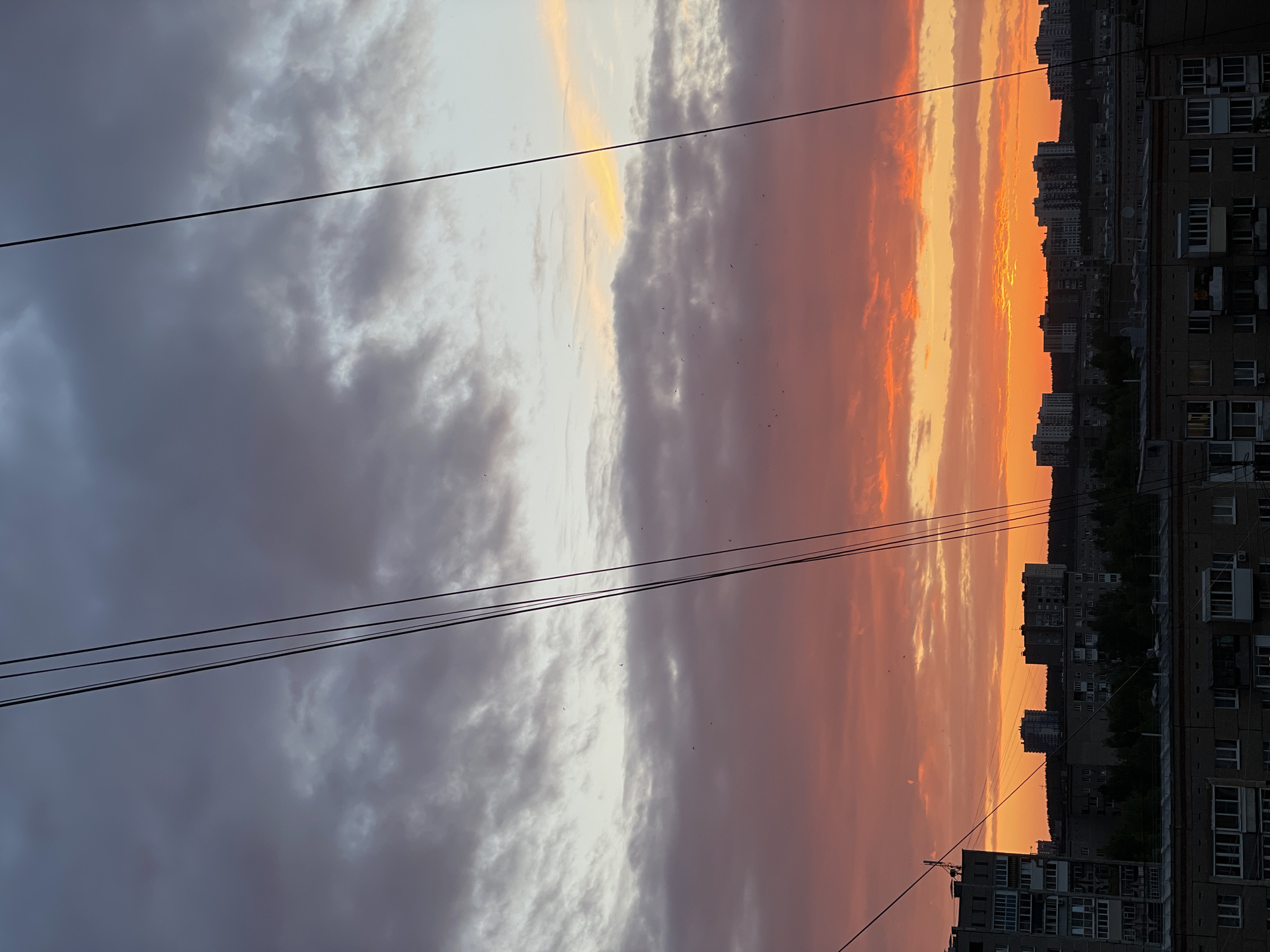 The image captures a dramatic sunset over the cityscape of Izhevsk, Russia. The sky is a vibrant spectacle of colors, transitioning from deep purples and grays at the top to fiery oranges and reds near the horizon. Wispy clouds are scattered across the sky, catching the last rays of the sun. Dominating the foreground are several thick, black power lines, starkly silhouetted against the colorful sky. Below the sky, a dense urban landscape unfolds. Multiple apartment buildings, varying in height and architectural style, stretch across the frame. Some buildings appear older, constructed of concrete and brick, while others are more modern high-rises. Trees and vegetation are visible between the buildings, suggesting a mix of residential and green spaces. The overall scene depicts a common urban vista at dusk, where natural beauty meets man-made structures. There are no people or active events visible in the image.