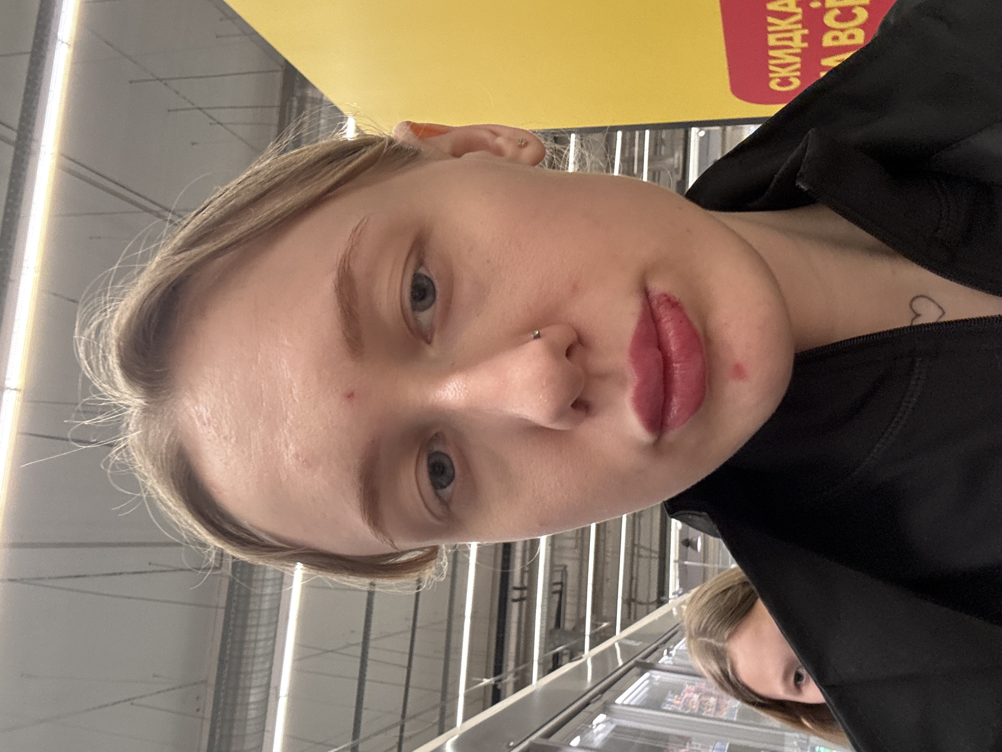 A selfie of a young woman with light skin, blonde hair pulled back, and a nose piercing, taken indoors in a commercial setting. She's wearing a black garment, possibly a jacket, with a small heart tattoo visible on her neck. Behind her are rows of refrigerated display cases, suggesting a supermarket or grocery store. Overhead, industrial lighting fixtures illuminate the scene. A yellow and red sign is partially visible in the background. The image does not confirm that the location is Rostov-on-Don, Russia.