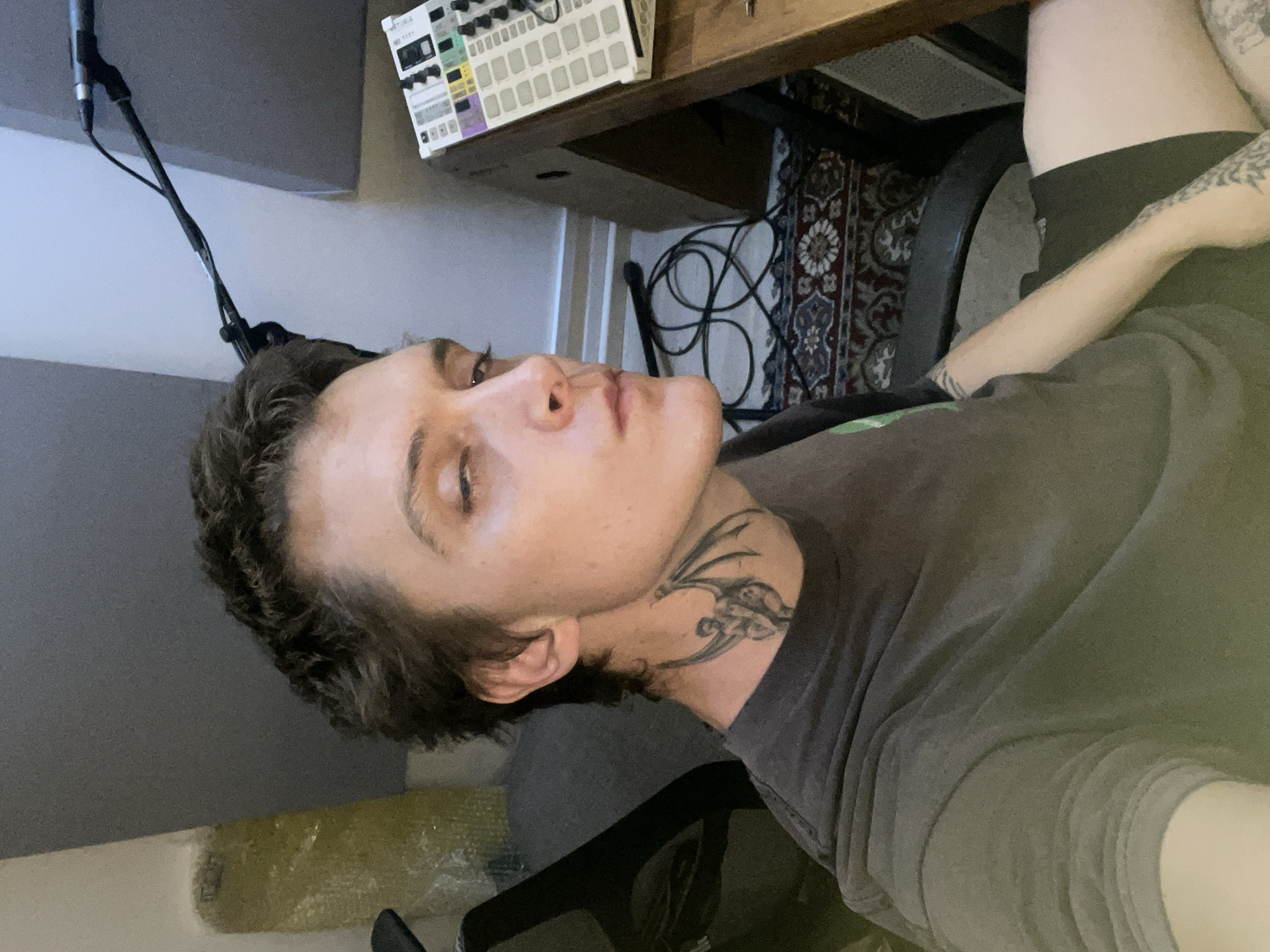 An indoor, horizontally-oriented image (rotated 90 degrees counter-clockwise) depicts a young individual, likely male, with short, dark, curly hair and light skin. The person is wearing a dark grey t-shirt and has a distinctive tribal-style tattoo on their neck. Their eyes are partially closed, and they are looking upwards and slightly away from the camera, giving a contemplative or relaxed impression.
The setting appears to be a home music studio or creative workspace. To the left of the subject's head, a black microphone stand is visible, extending upwards towards what looks like dark grey acoustic paneling on the wall. In the background, behind the person's head, a wooden desk holds a white electronic music production device or synthesizer with colorful knobs and buttons, bearing the visible brand name "ARTURIA." Below the desk, a patterned rug with intricate designs covers the floor, with loose cables coiled on it. A dark-colored office chair is partially visible under the desk, and a bare leg with tattoos and an arm are seen on the right side of the frame. The lighting is ambient, suggesting an indoor environment. The scene captures a moment of rest or introspection within a creative space.