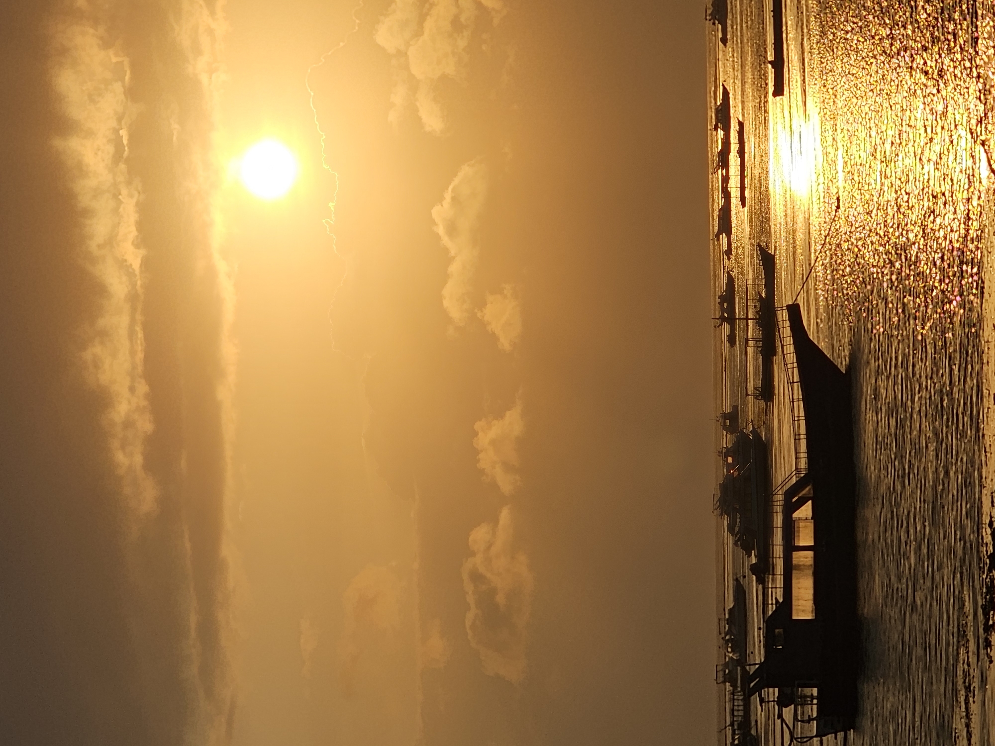 A serene sunset over a body of water is depicted, likely in Denpasar, Indonesia. The sky is a vibrant gradient of orange and yellow, with the sun a bright orb positioned in the upper right quadrant, partially obscured by soft, scattered clouds. The water's surface reflects the golden hues of the sky, creating a shimmering, textured effect.
Several boats are scattered across the water. In the foreground, a dark silhouette of a boat with visible windows is prominent. Further out, more boats, varying in size and type, are anchored or drifting. Some appear to be leisure crafts, while others might be fishing vessels. The overall atmosphere is peaceful and tranquil, characteristic of a coastal scene at dusk. There are no people visible in the image. The environment suggests a coastal area, possibly a harbor or a bay, typical of locations near Denpasar where water activities and boat traffic are common.