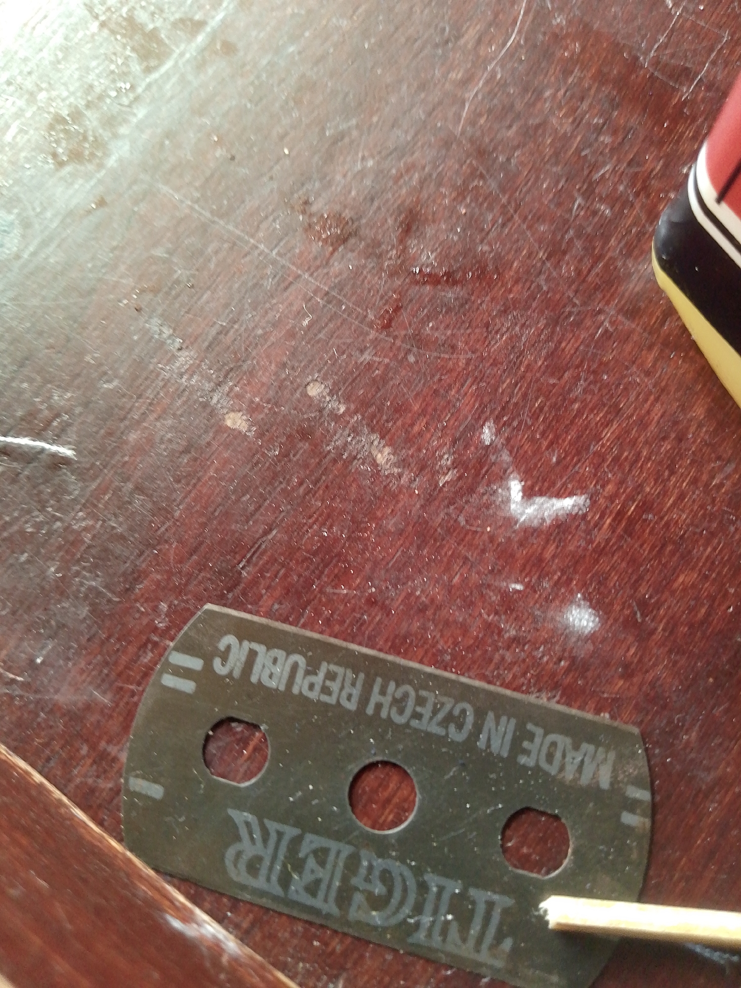 The image shows a close-up, overhead view of a razor blade resting on a dark wooden surface. The razor blade is dark grey with white text and markings. The text reads "MADE IN CZECH REPUBLIC" and what appears to be a brand name starting with "TIGER". There are three circular holes in the blade. A small, light-colored object, possibly a toothpick or a piece of straw, is partially visible extending from the right side of the image, touching the razor blade near one of the holes. The wooden surface is scratched and appears to have some minor smudges or debris on it. To the upper right of the razor blade, a portion of a red and yellow object is visible, partially out of frame. The scene is lit from above, creating some glare and reflections on the wooden surface. There is no indication of the city of Jauro Yinu, Nigeria, or any other specific location within the image.