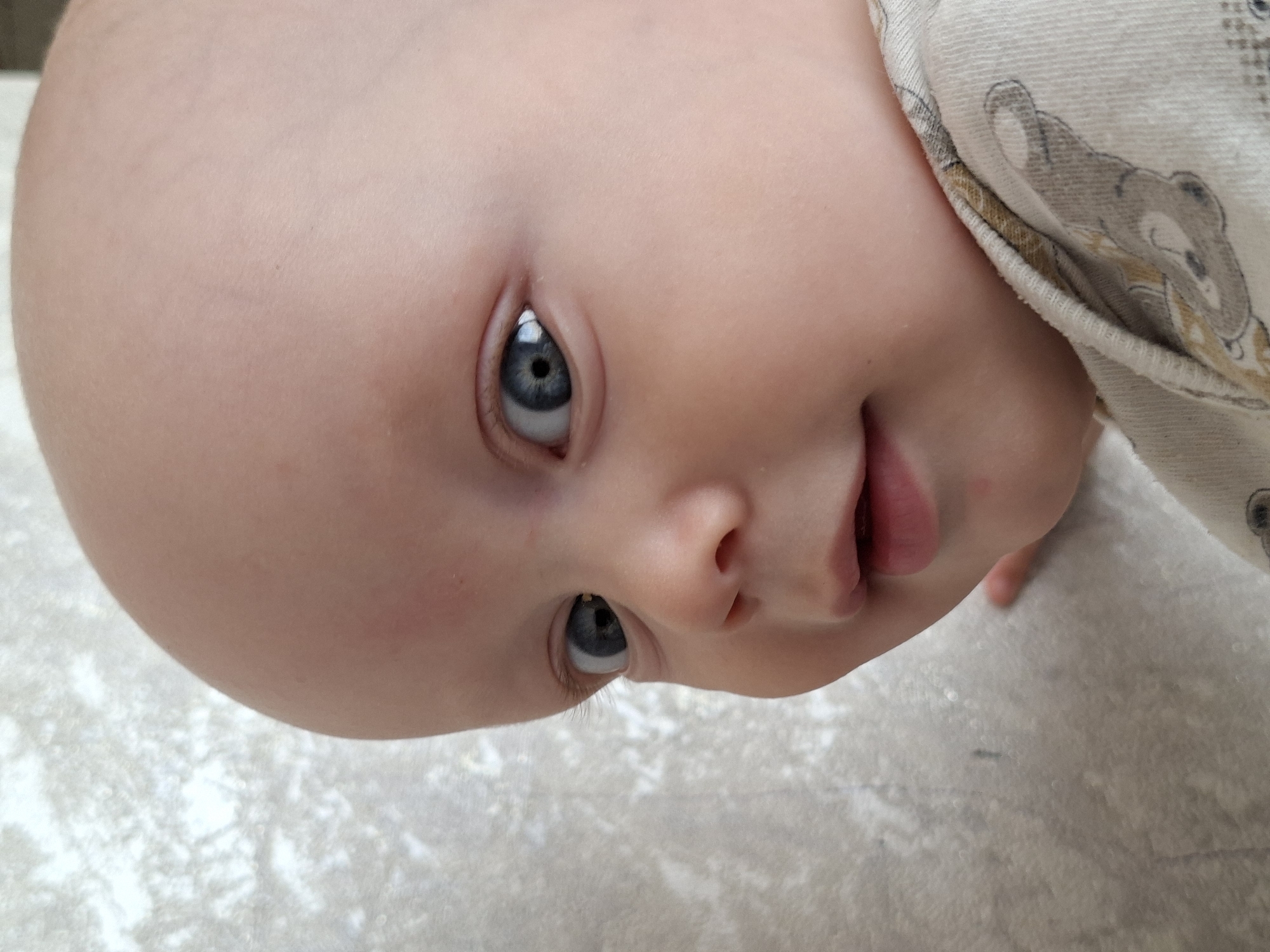 A close-up, eye-level, vertical, outdoor medium shot captures a baby's face. The baby has light skin, a bald head, and striking blue eyes. The left eye is fully visible, looking directly at the camera, while the right eye is partially obscured. The baby's mouth is slightly open, revealing the lower lip. The baby is wearing a light-colored onesie with a gray and yellow pattern of teddy bears. The background is a textured, light gray surface, possibly a wall or blanket. The lighting is soft and natural, highlighting the baby's features. The image is in focus, with a shallow depth of field that blurs the background.