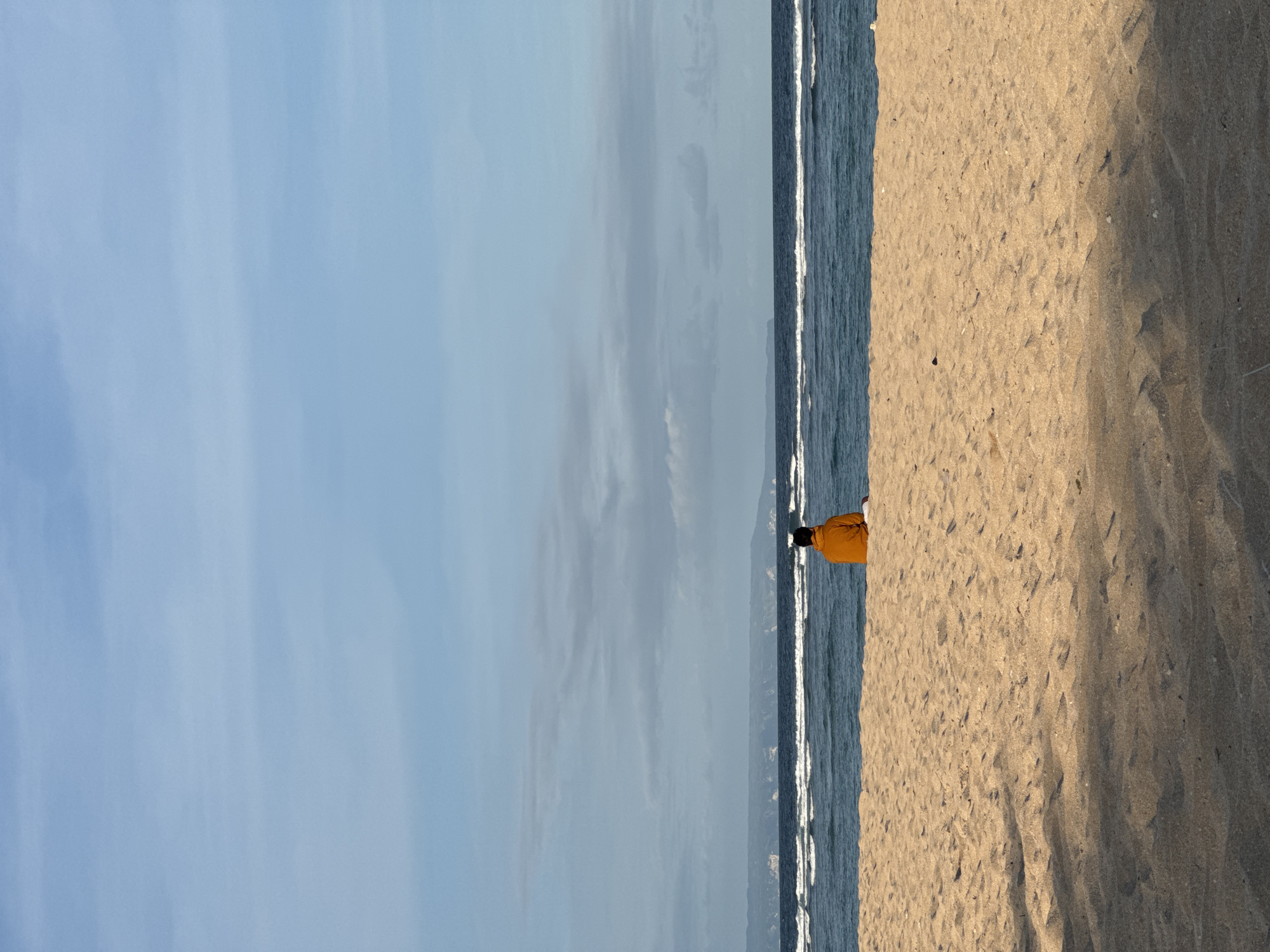A vertical shot depicts a beach scene. The sky dominates the left portion of the frame, exhibiting light blue hues with subtle cloud formations. Adjacent to the sky is the ocean, characterized by gentle waves cresting with white foam. To the right of the ocean is the sandy beach, occupying the remaining portion of the image. A solitary figure, clad in an orange shirt, stands near the shoreline on the beach. The image is most likely taken in Kuta Selatan, Indonesia.