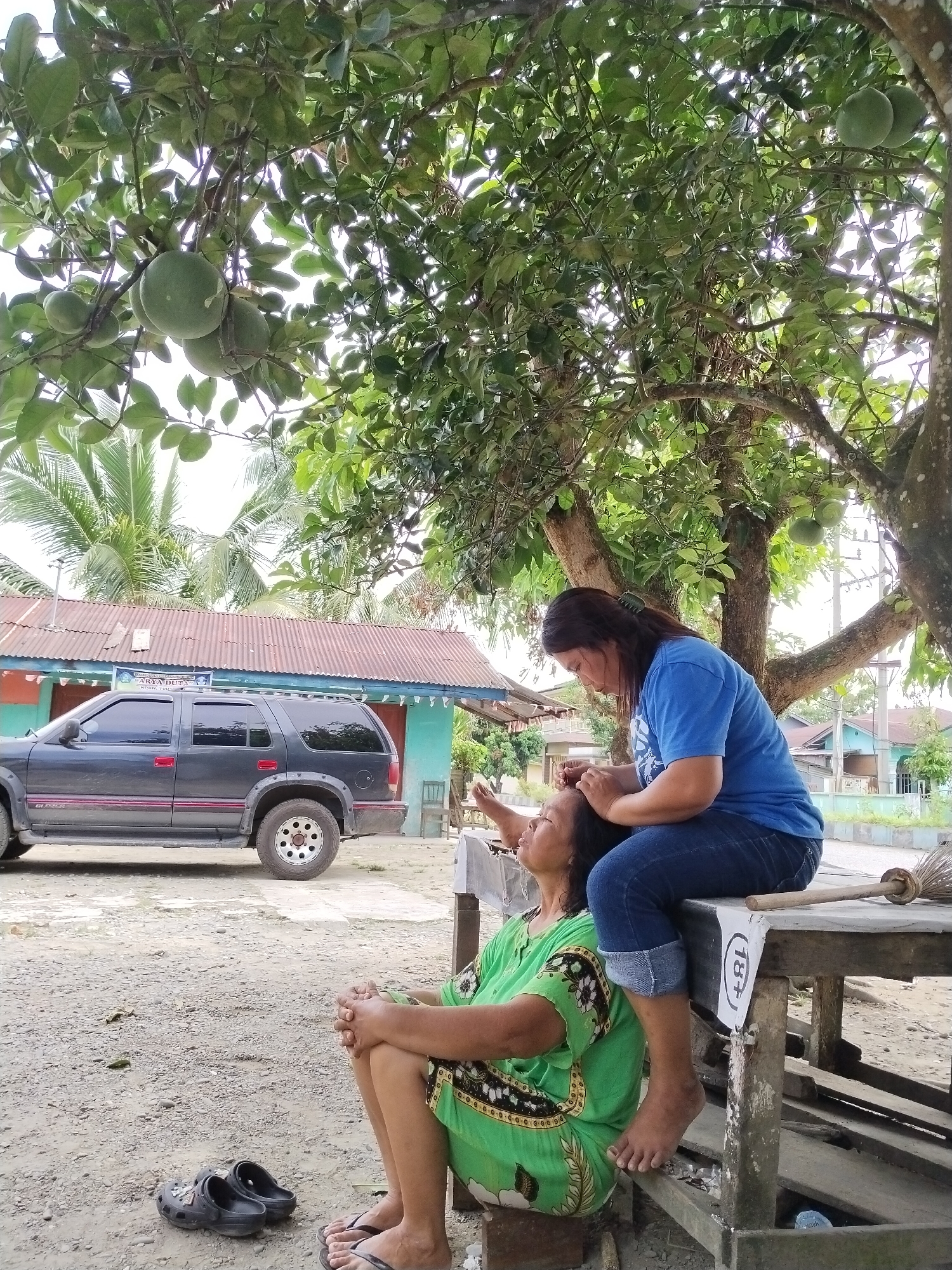 In Sawit Seberang, Indonesia, under the dappled shade of a large tree laden with green citrus fruits, two women are engaged in an outdoor activity. One woman, wearing a blue t-shirt and jeans, is seated on a wooden table, leaning forward to give a head massage to another woman. The woman receiving the massage is seated below, dressed in a vibrant green dress adorned with a floral pattern, her eyes closed and head tilted back in apparent relaxation. Their interaction is intimate and caring. In the background, a dark-colored SUV is parked on a dirt lot, and a simple corrugated-roof building with painted walls is visible. The environment suggests a rural or semi-rural setting, characterized by the natural elements of the tree and foliage, and the modest infrastructure. The overall scene conveys a sense of peace and community.