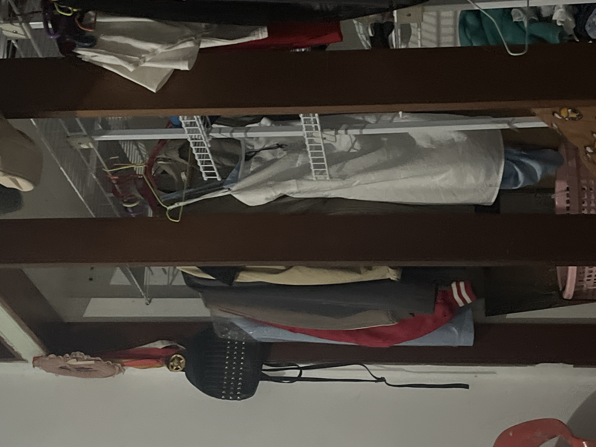 The image depicts an interior view of a closet or wardrobe. Hanging on the left side, from a hook, is a black studded backpack with a dangling strap. Above the backpack, a circular, decorative ribbon with gold accents is visible. To the right of the backpack, several garments are hanging on a rod, including a red and white striped sleeve, a blue garment, a grey jacket, and a tan jacket.
Further to the right, a white garment, possibly a dress or apron, hangs from a rack with multiple shelves. Several hangers, some colored, are visible in the background. On a higher shelf, a beige cap or hat is placed. To the far right, more clothing items are visible on hangers, including white t-shirts and dark-colored garments. A red item is partially visible on a shelf. At the bottom right, the edge of a teal garment and a white shelf are visible.
The scene appears to be in a residential setting. The overall lighting is dim. There are no people visible in the image, and no actions are being performed. The image does not provide any specific visual cues to place it in Santa Ana, Venezuela.