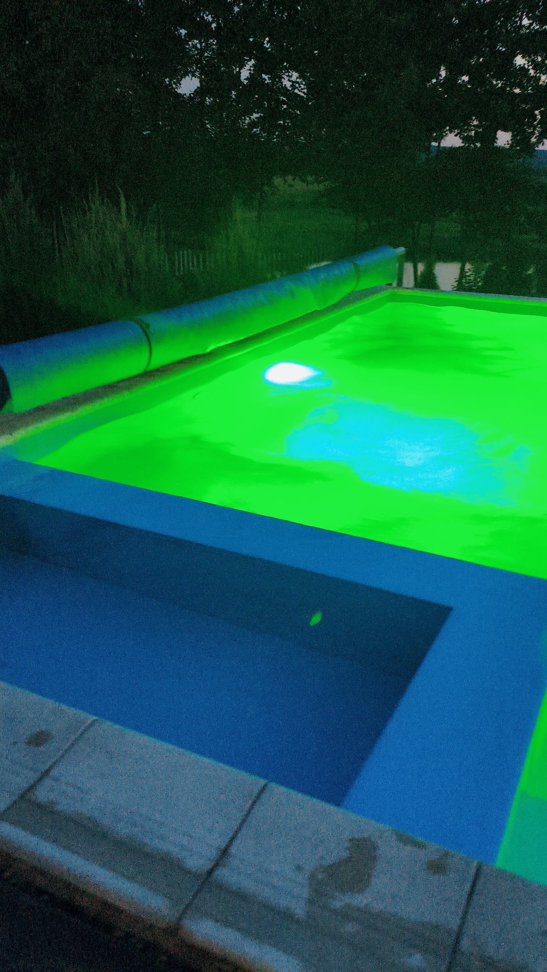 The image shows an outdoor swimming pool at night, illuminated by green lighting. The pool is rectangular and appears to be partially covered by a rolled-up cover. The water is a vibrant green color, with a bright white reflection on the surface. The surrounding area is dark, with trees and vegetation visible in the background. The pool deck is made of gray paving stones. There are no people visible in the image. The location is stated to be in Bezliudivka, Ukraine.